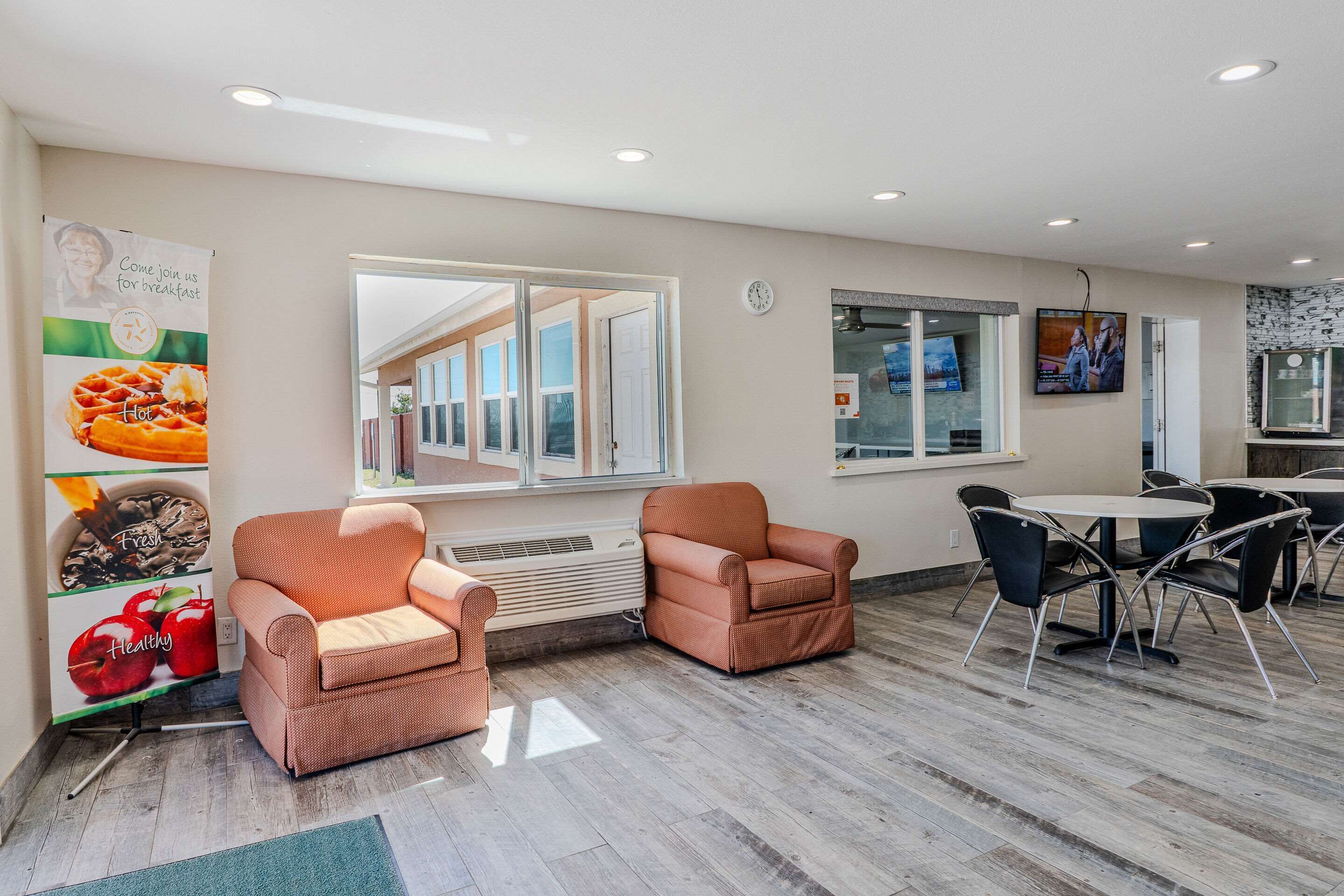 A modern hotel lobby with two orange armchairs, a round table with chairs, a large window, and a poster advertising breakfast.