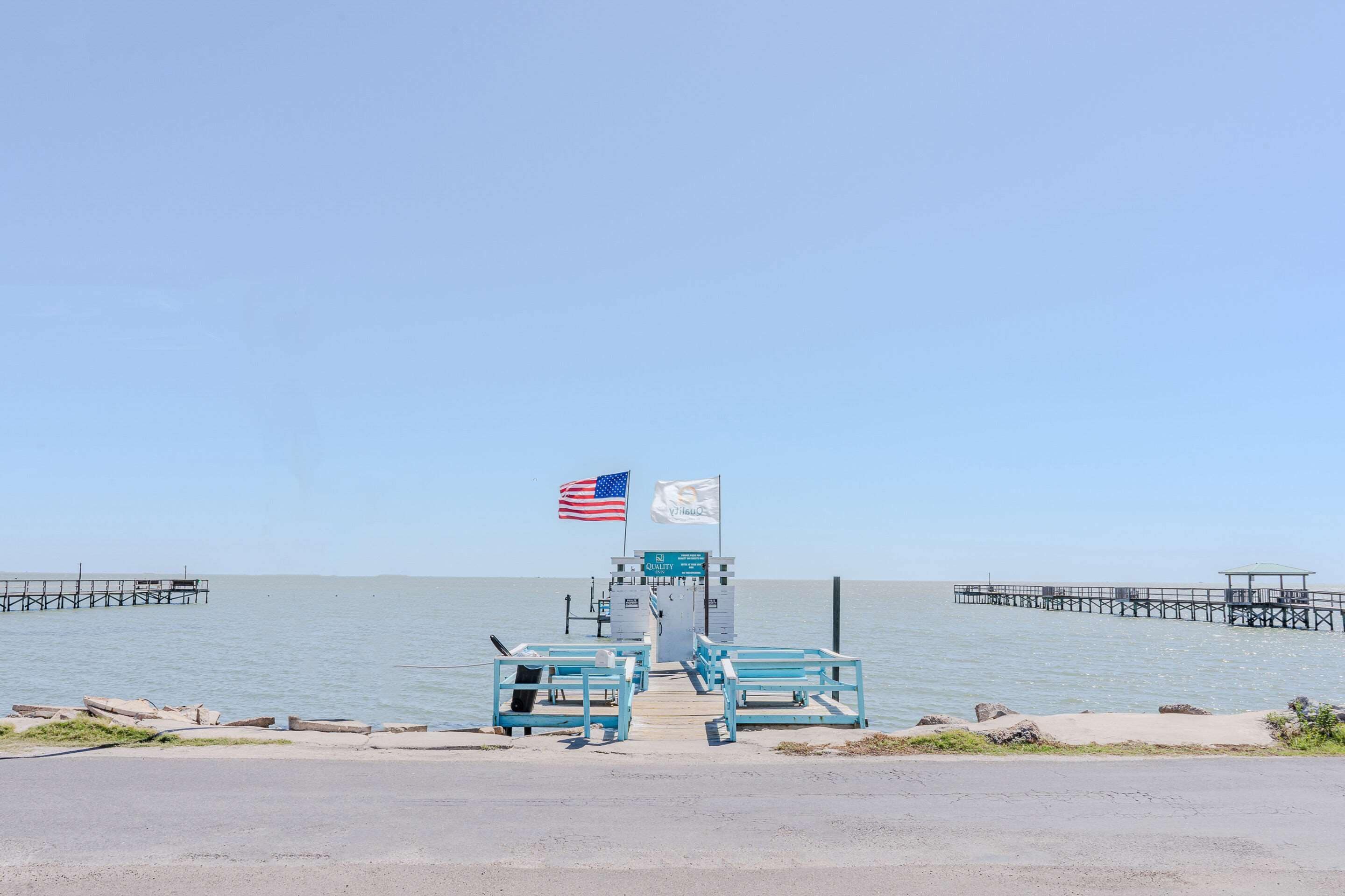 A pier with a small building and two flags, a road leading to it, and calm water in the background.