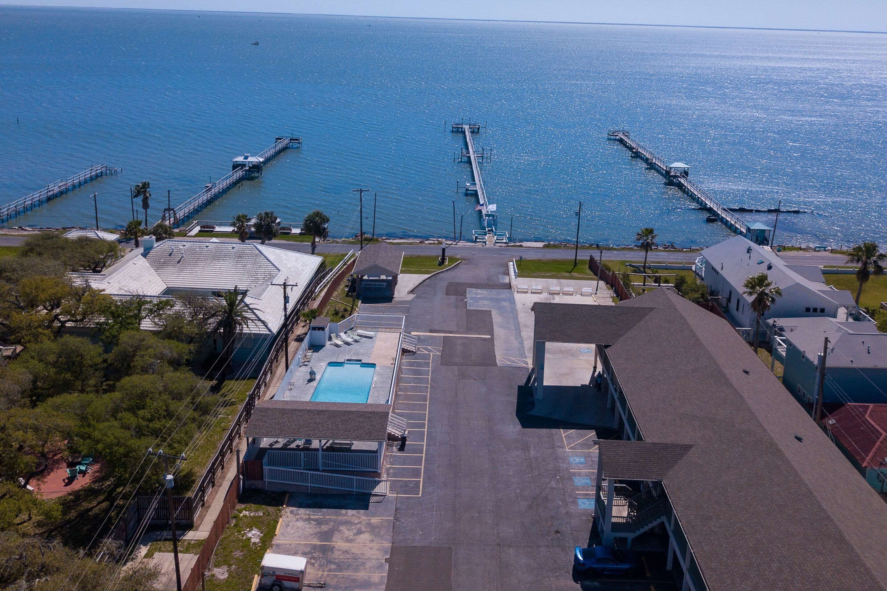 An aerial view of a coastal area with a long pier extending into the sea, a swimming pool, and adjacent buildings.