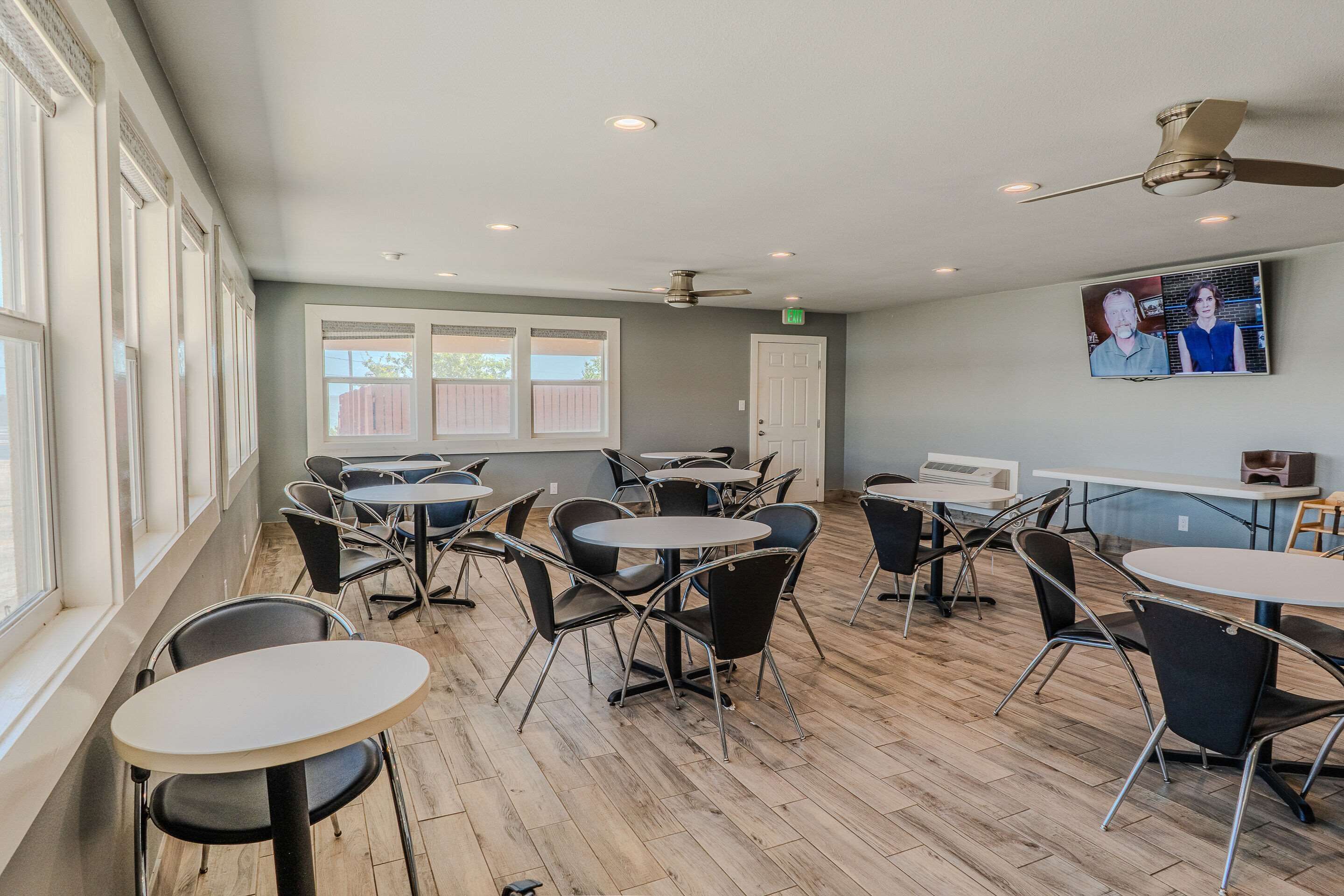 A spacious room with multiple round tables and chairs, large windows, and a ceiling fan.