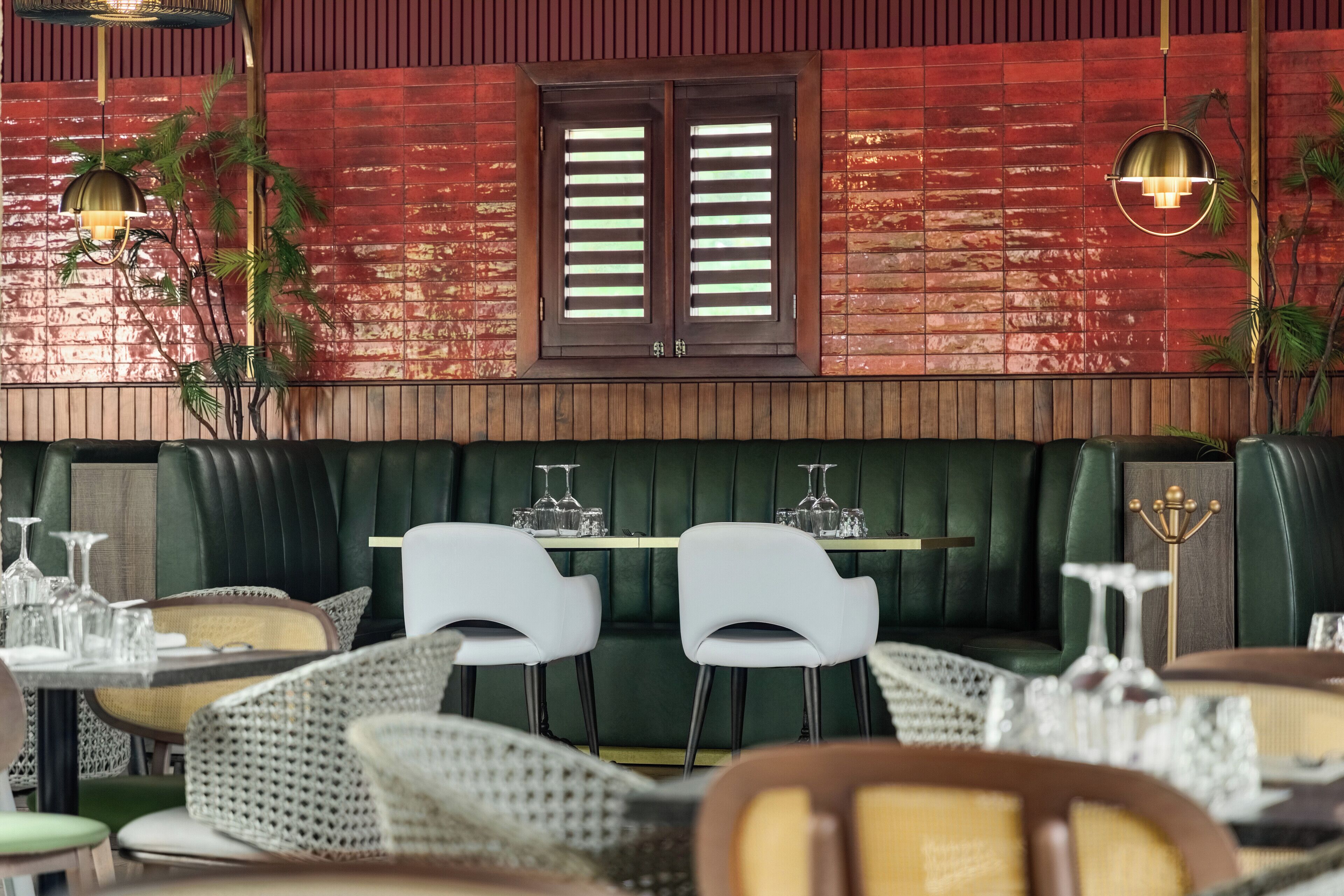 Un restaurant avec un coin salon doté de banquettes vertes, de chaises blanches et d’un mur en briques agrémenté d’une fenêtre.