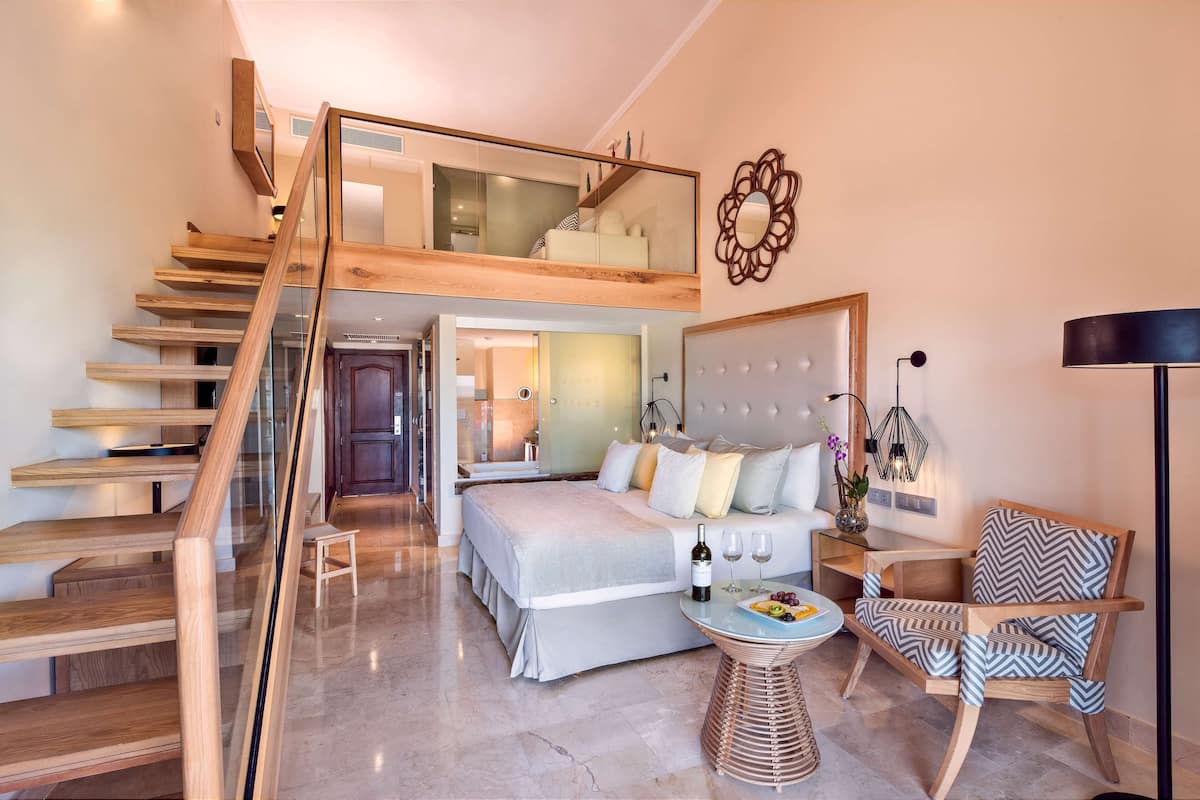 Een moderne slaapkamer met een groot bed, een glazen trap, een eethoek met tafel en stoelen, en een zithoek met een bank en een lamp.