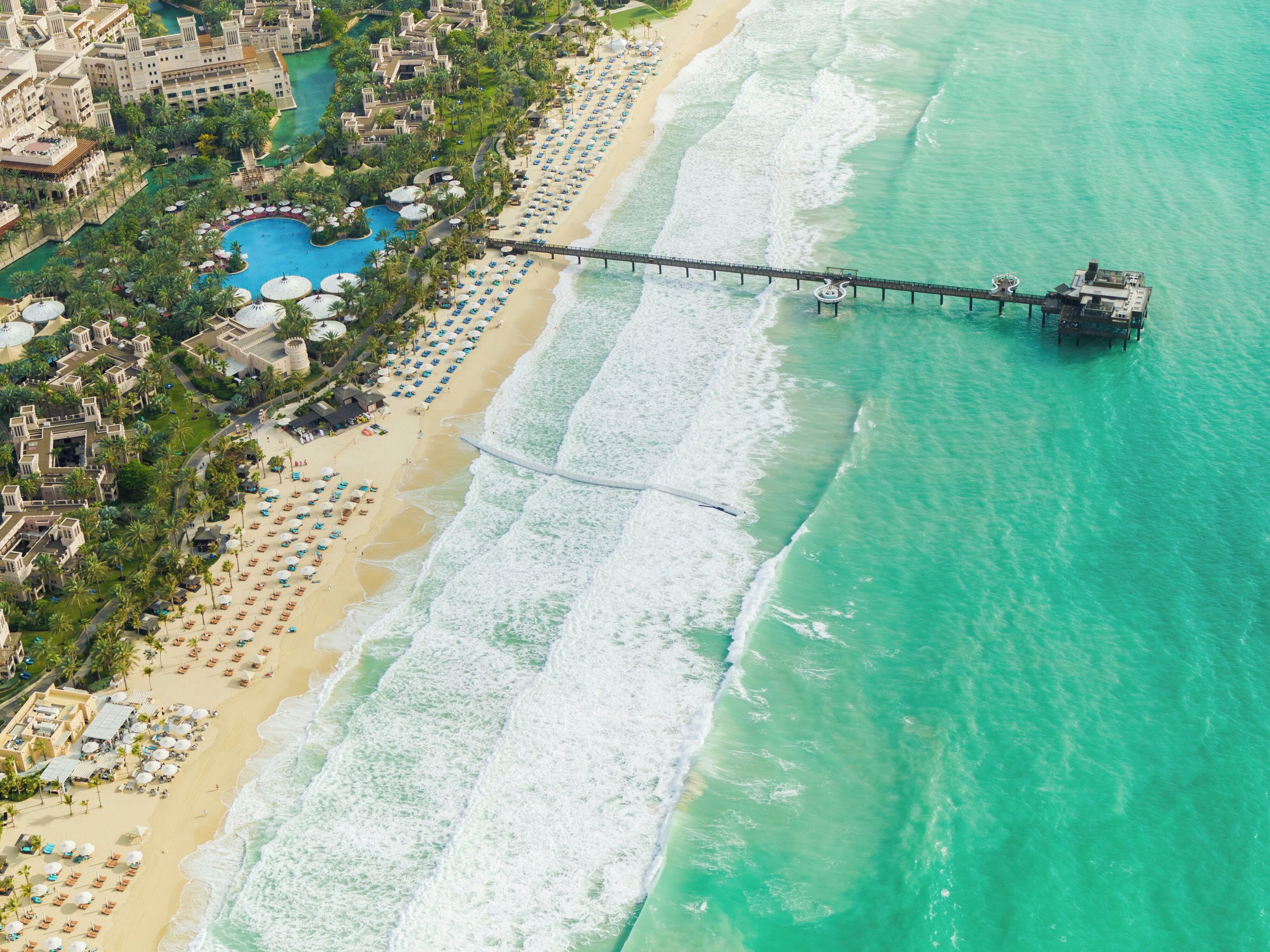 An aerial view of a resort with a long pier extending into turquoise waters, a sandy beach lined with umbrellas, and a pool area.