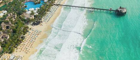 An aerial view of a resort with a long pier extending into turquoise waters, a sandy beach lined with umbrellas, and a pool area.