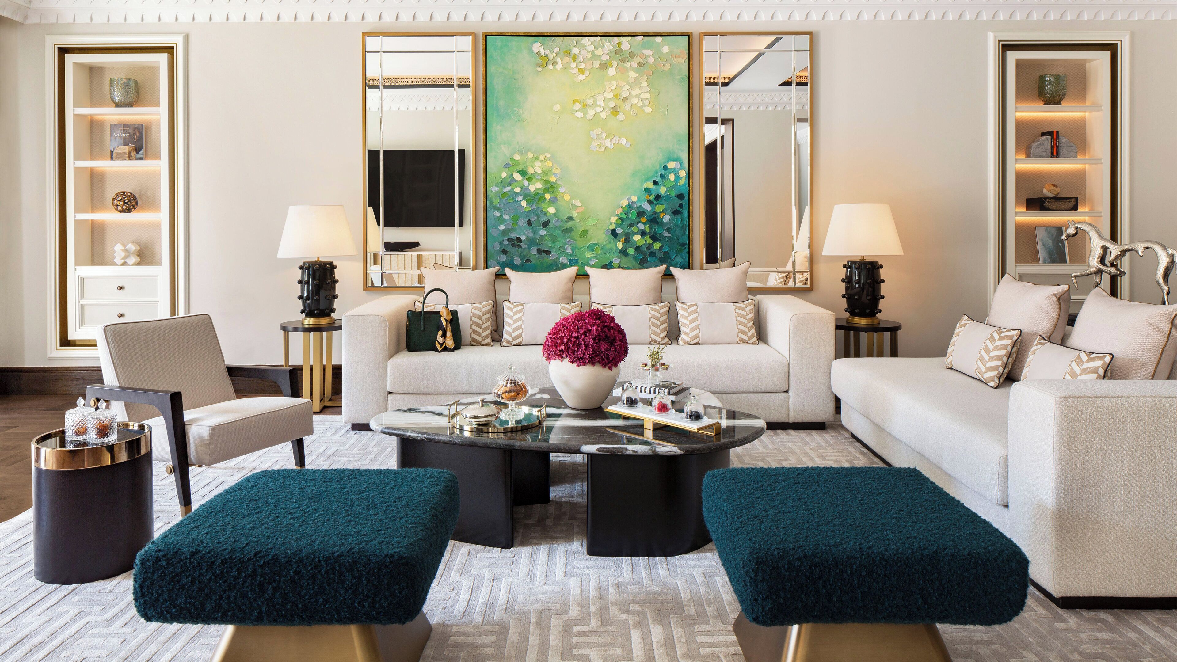 A modern living room with a sofa, two ottomans, a coffee table, and decorative items. There are two lamps, a vase with flowers, and a painting on the wall.