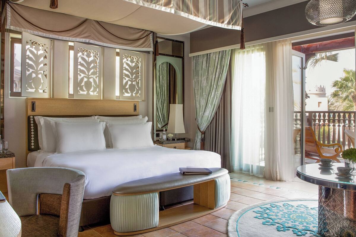 Una habitació d'hotel àmplia amb un llit gran, una zona de seients amb una taula i cadires, un balcó amb vistes i un sostre decoratiu.