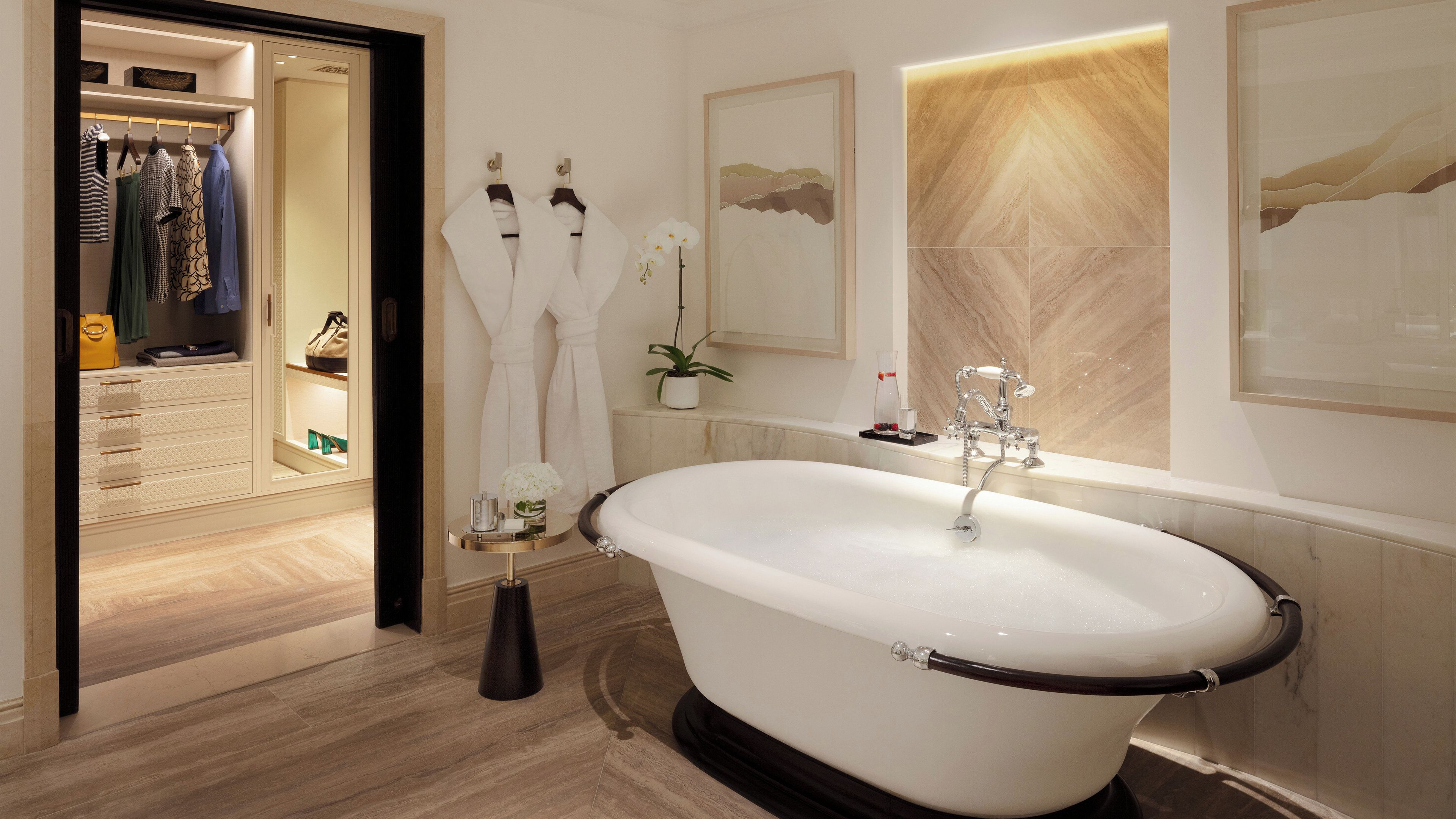 A modern bathroom with a freestanding bathtub, a walk-in closet with hanging clothes, and a large mirror with a decorative wall panel.