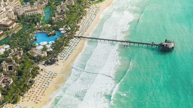 Eine Luftaufnahme eines Küstenresorts mit einer langen Pier, die sich in türkisfarbenes Wasser erstreckt, einem Sandstrand mit Reihen von Sonnenschirmen und einem Poolbereich, der von Palmen umgeben ist.