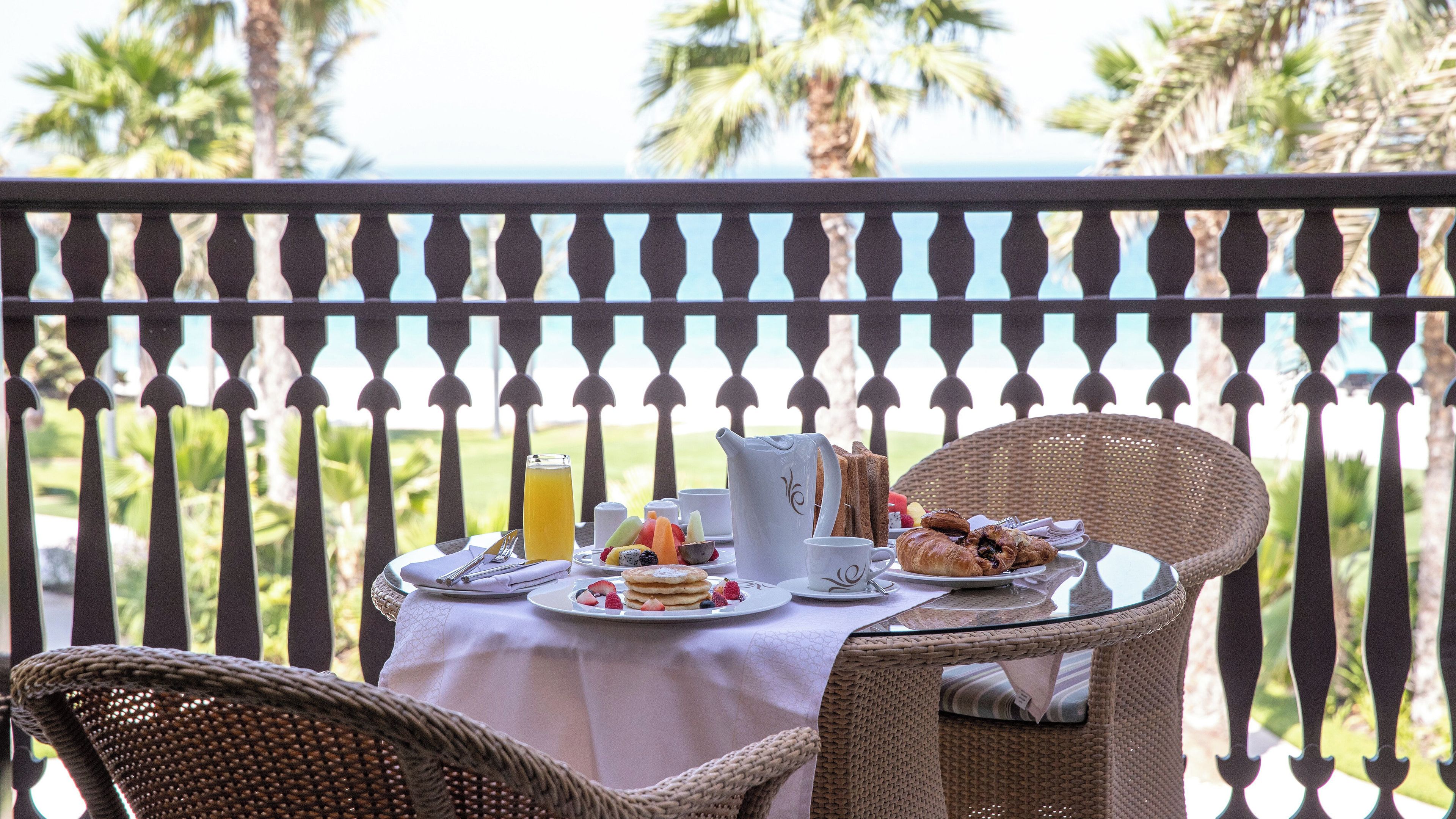 A dining table with breakfast items and a pitcher of orange juice on a balcony with a view of palm trees and the ocean.
