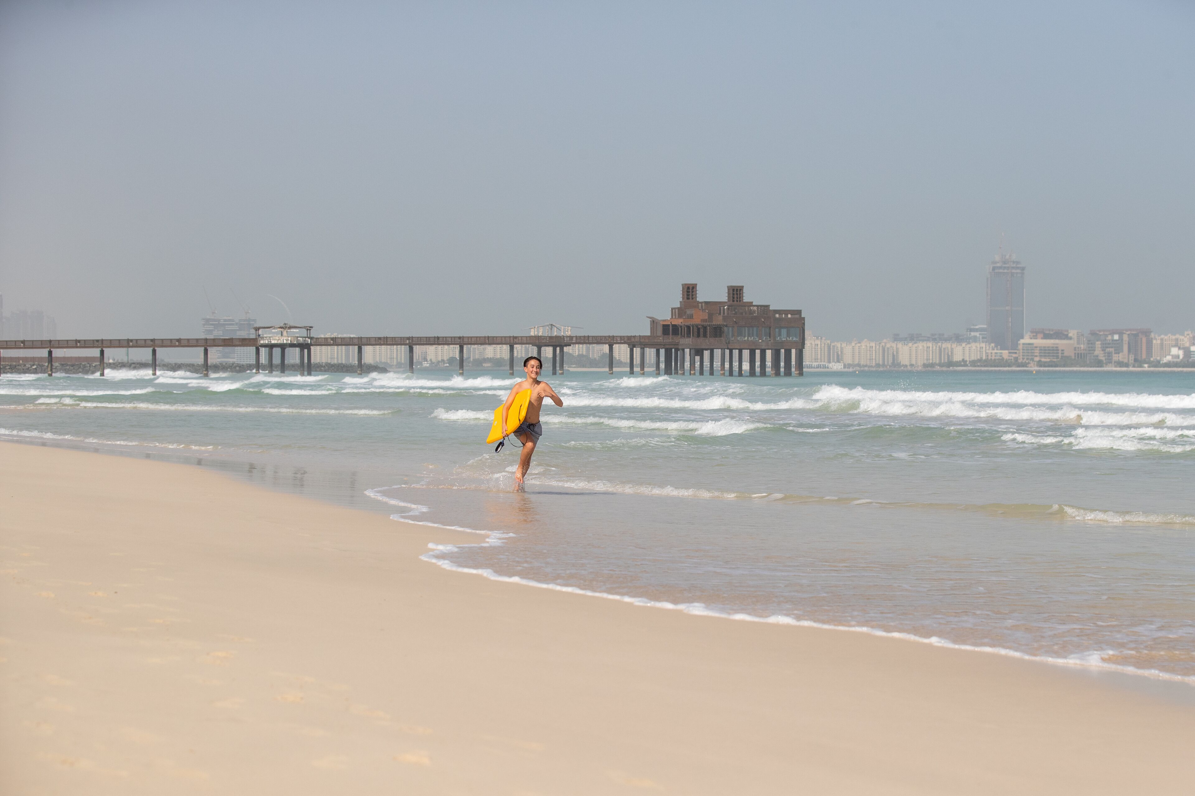 Una playa con un muelle que se adentra en el océano, un horizonte urbano de fondo y una persona llevando una tabla de surf amarilla.