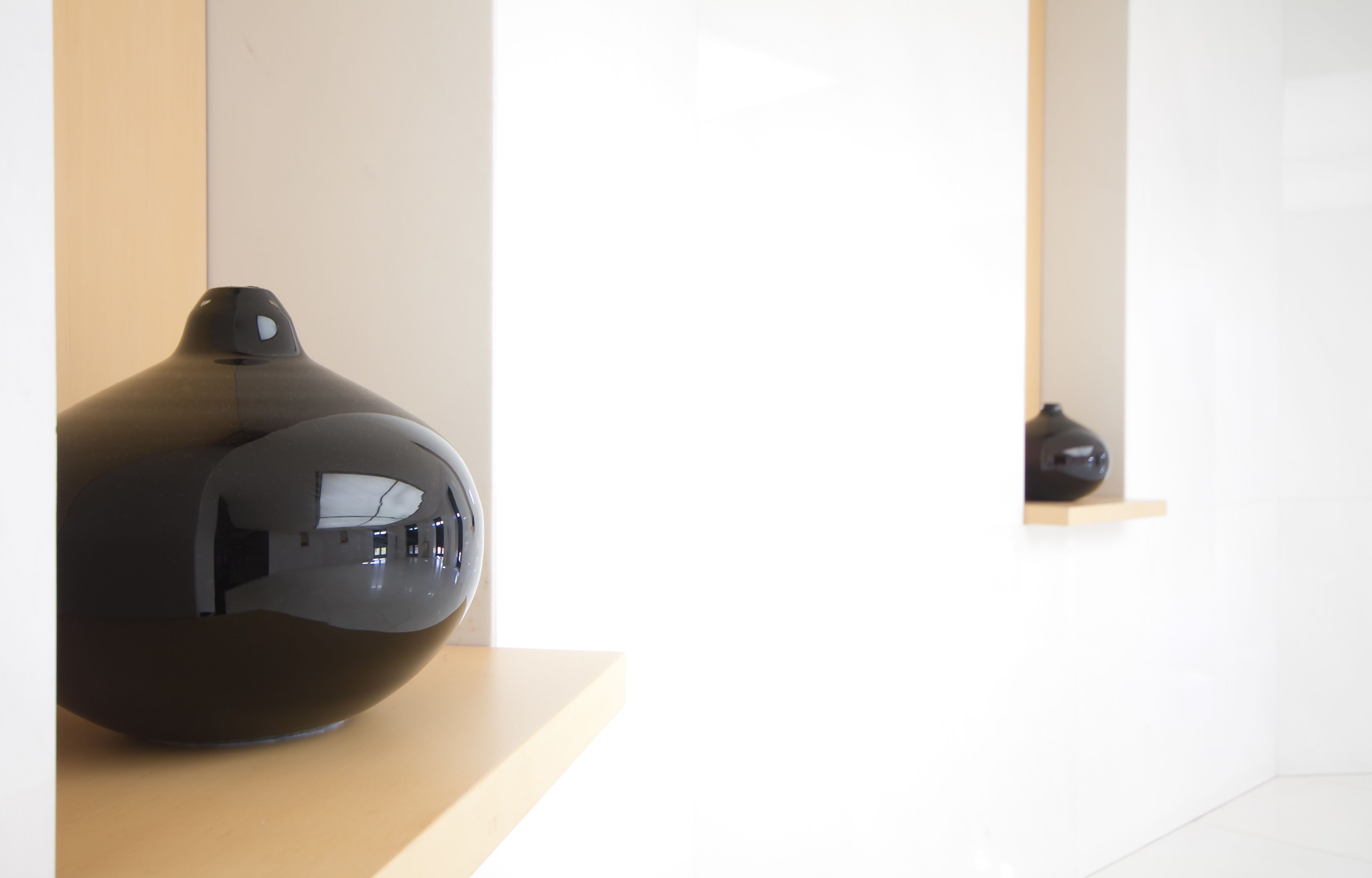 Habitación moderna y minimalista con un objeto negro esférico sobre una superficie de madera, una pared blanca y un estante con otro objeto negro.