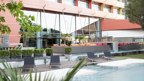A modern outdoor pool area with lounge chairs and seating, surrounded by a building with large windows and a red roof. - Hotel Puerta América (Madrid)