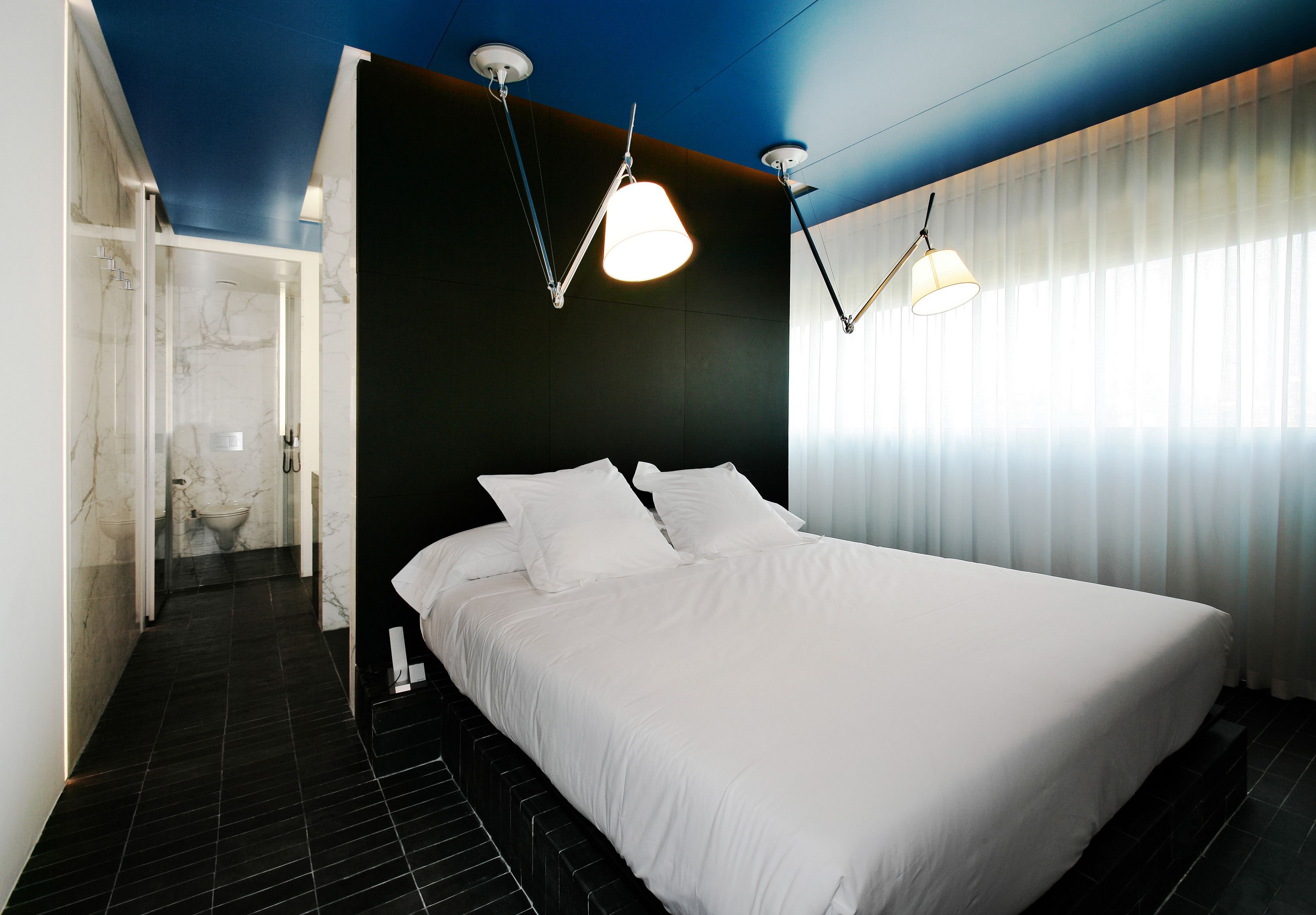 A modern hotel room with a large bed, a walk-in shower, and a view of the outdoors through sheer curtains.