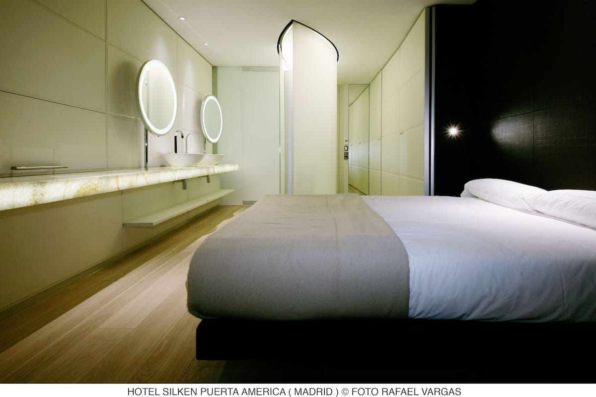 A modern hotel room with a large bed, a vanity with two mirrors, and a dark accent wall.