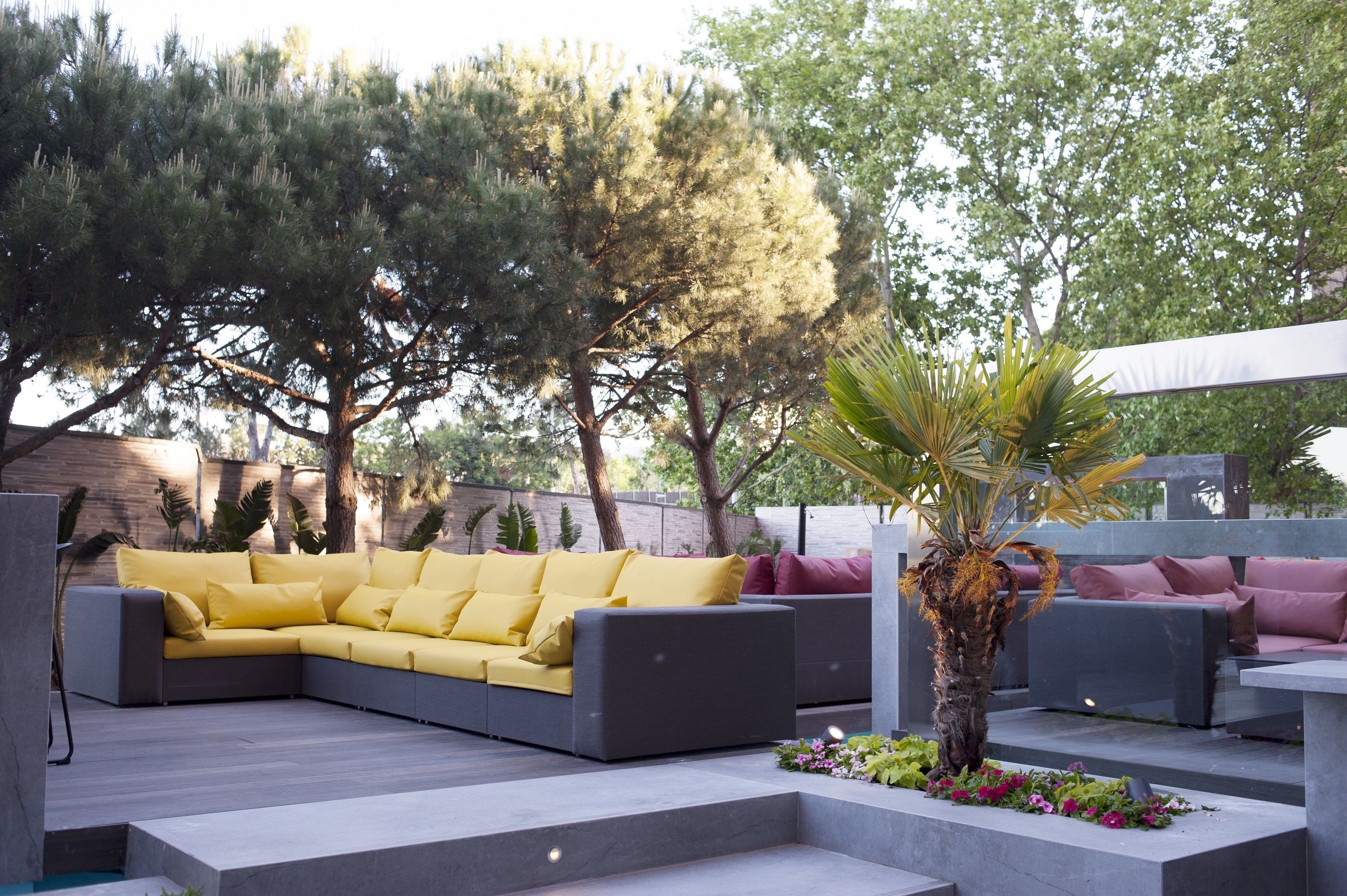 Un espacio al aire libre moderno con zona de estar, un sofá gris y cojines amarillos, rodeado de vegetación y árboles.