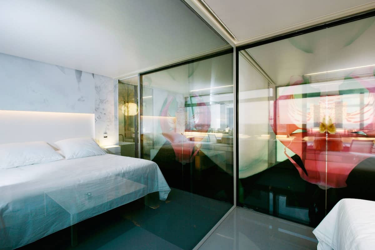 A modern hotel room with a bed, a glass partition, and a view of the cityscape.