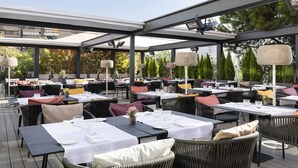 An outdoor dining area with tables and chairs, covered by a roof, and surrounded by greenery. - Hotel Puerta América (Madrid)