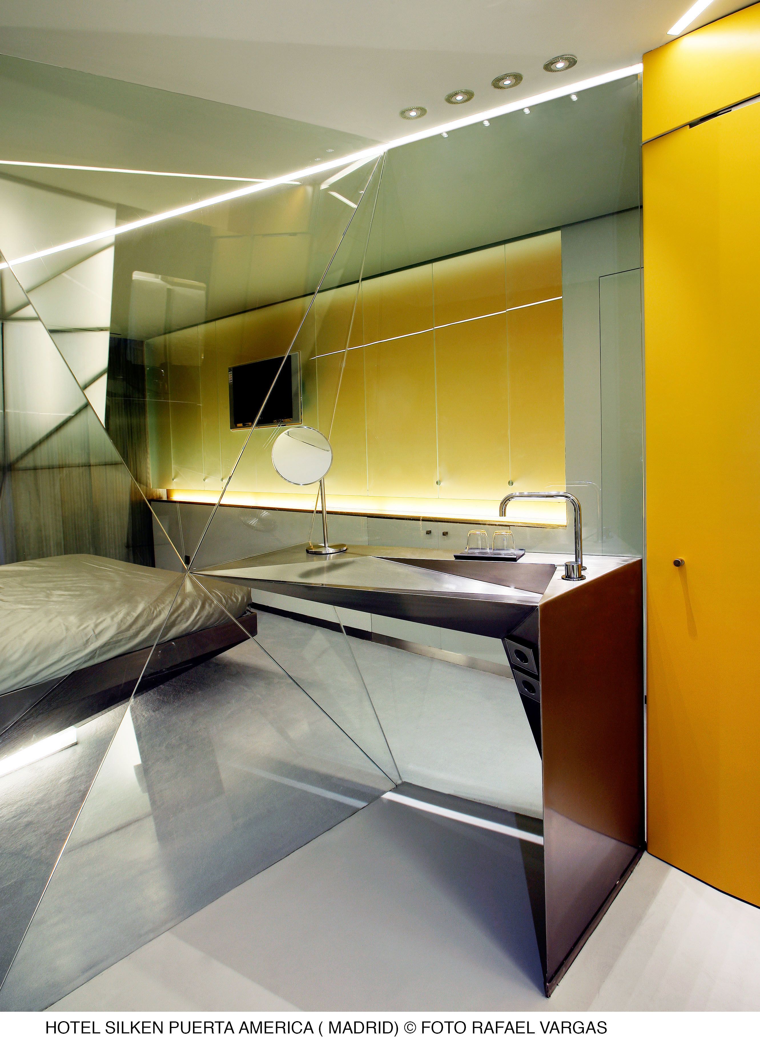 A modern hotel room with a glass partition, a bed, a mirror, and a sink.