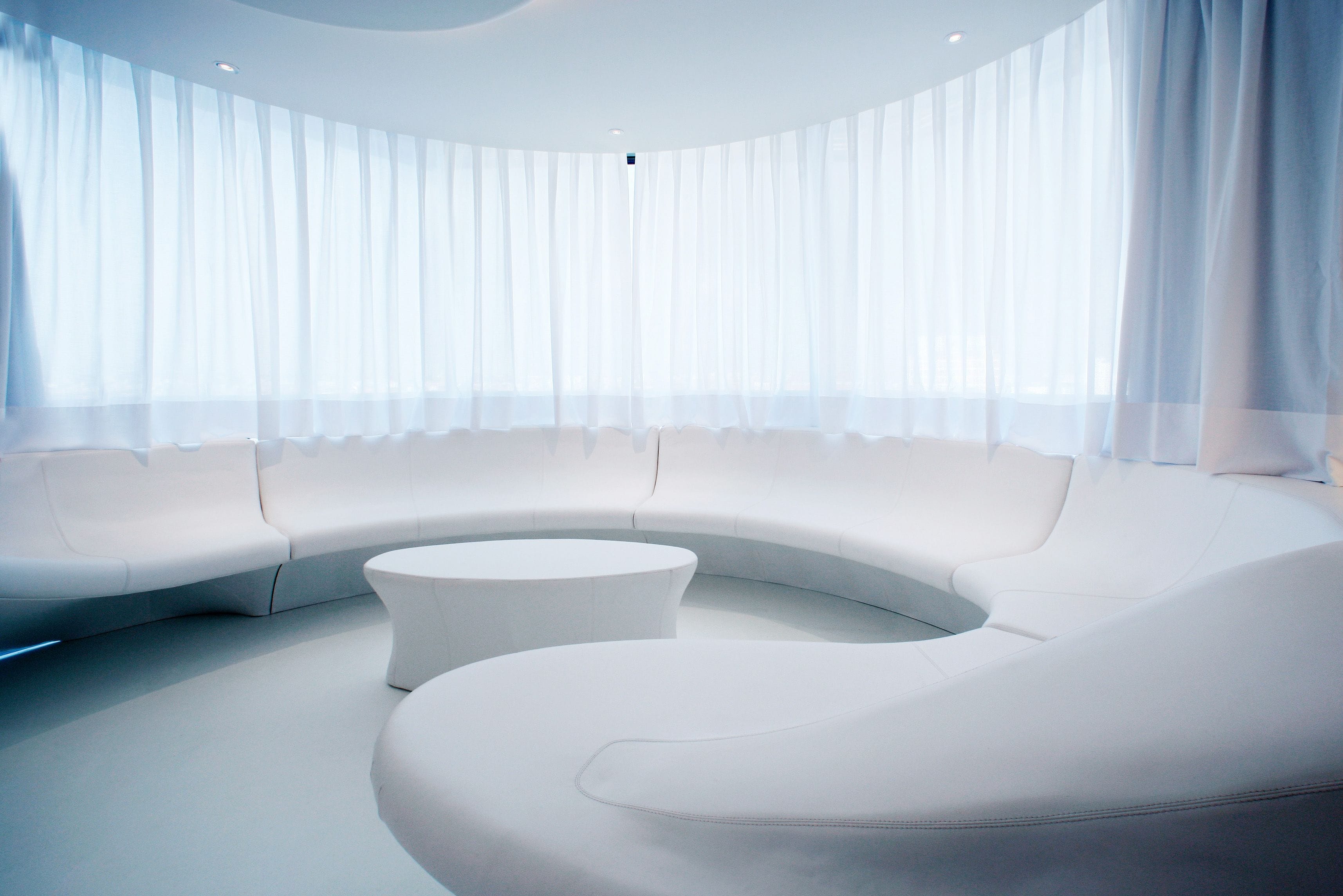 Ein modernes, gebogenes weißes Sofa und ein runder weißer Hocker in einem großzügigen Raum mit großen Fenstern und transparenten Vorhängen.