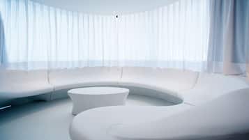 Ein modernes, gebogenes weißes Sofa und ein runder weißer Hocker in einem großzügigen Raum mit großen Fenstern und transparenten Vorhängen.