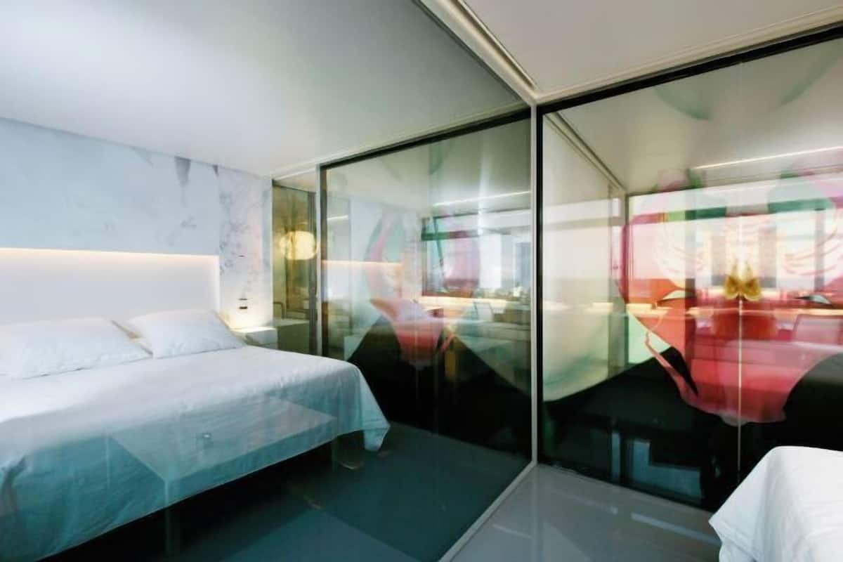 A modern hotel room with a bed, a glass partition, and a view of the cityscape.