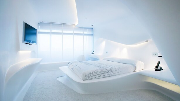 A modern bedroom with a curved bed, a flat-screen TV, and a large window. - Hotel Puerta América (Madrid)