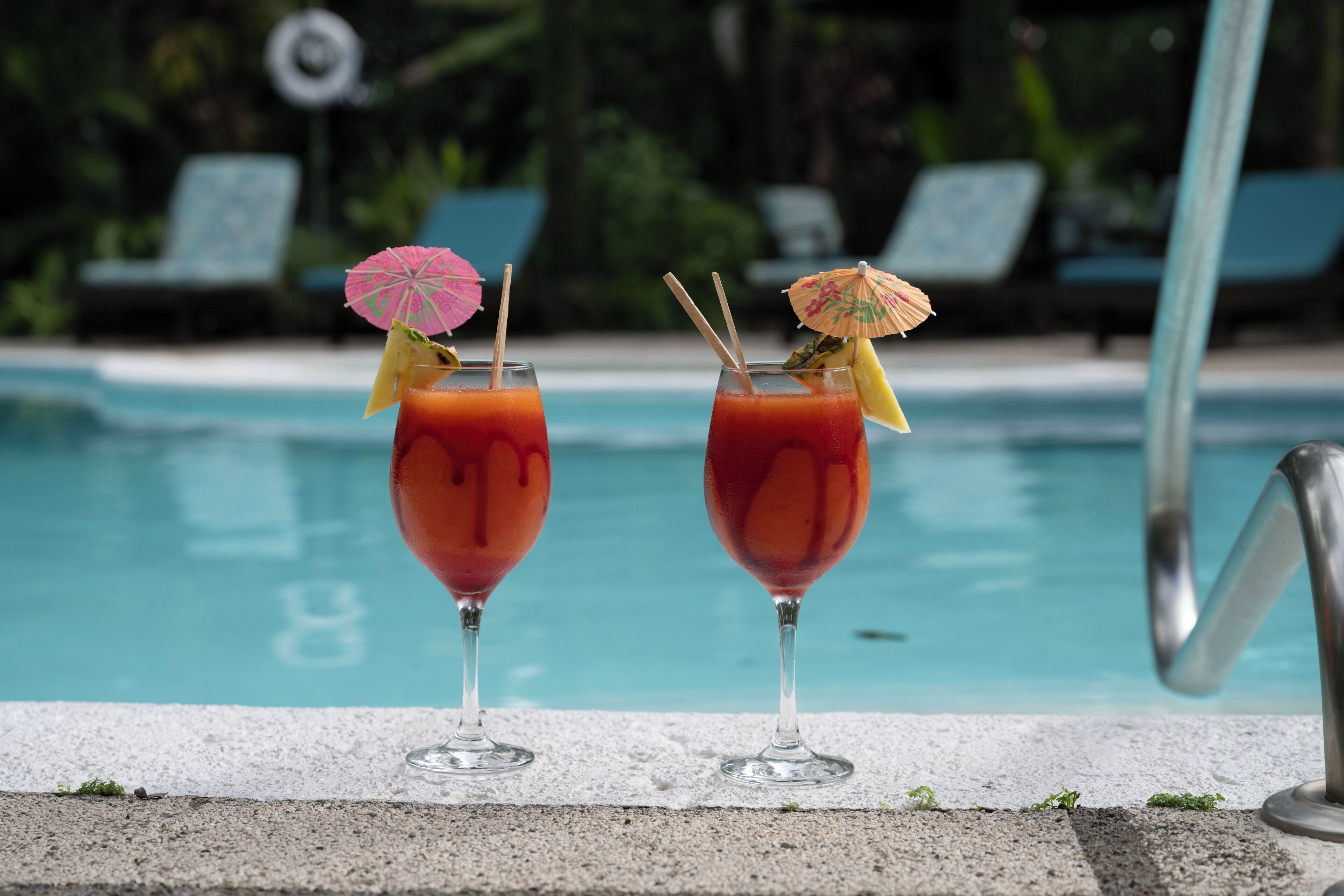 Two cocktails with umbrellas by a poolside, with lounge chairs and a pool in the background.