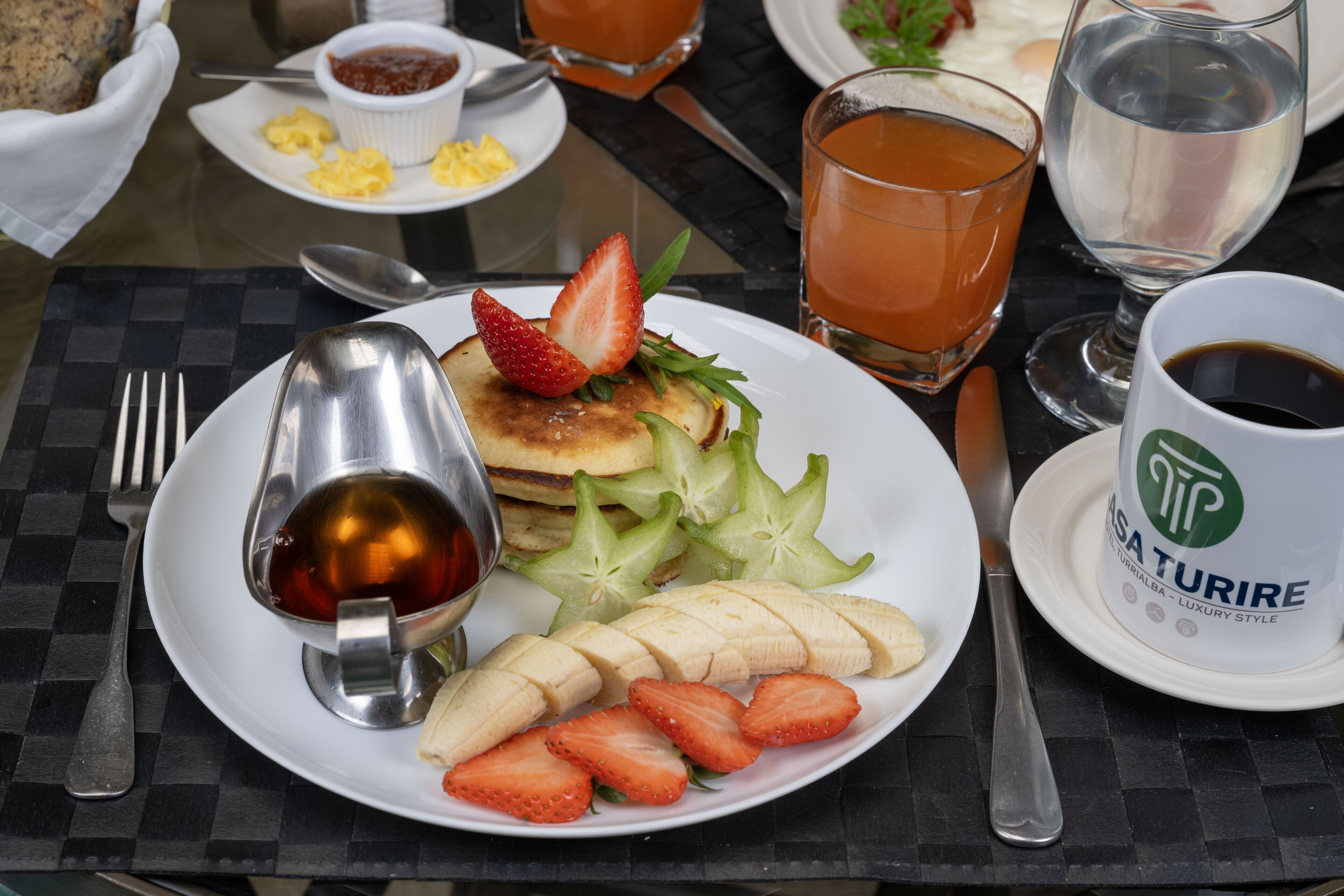 Een ontbijttafel met pannenkoeken, aardbeien en bananen op een bord, vergezeld van een kop koffie en een glas jus d'orange.