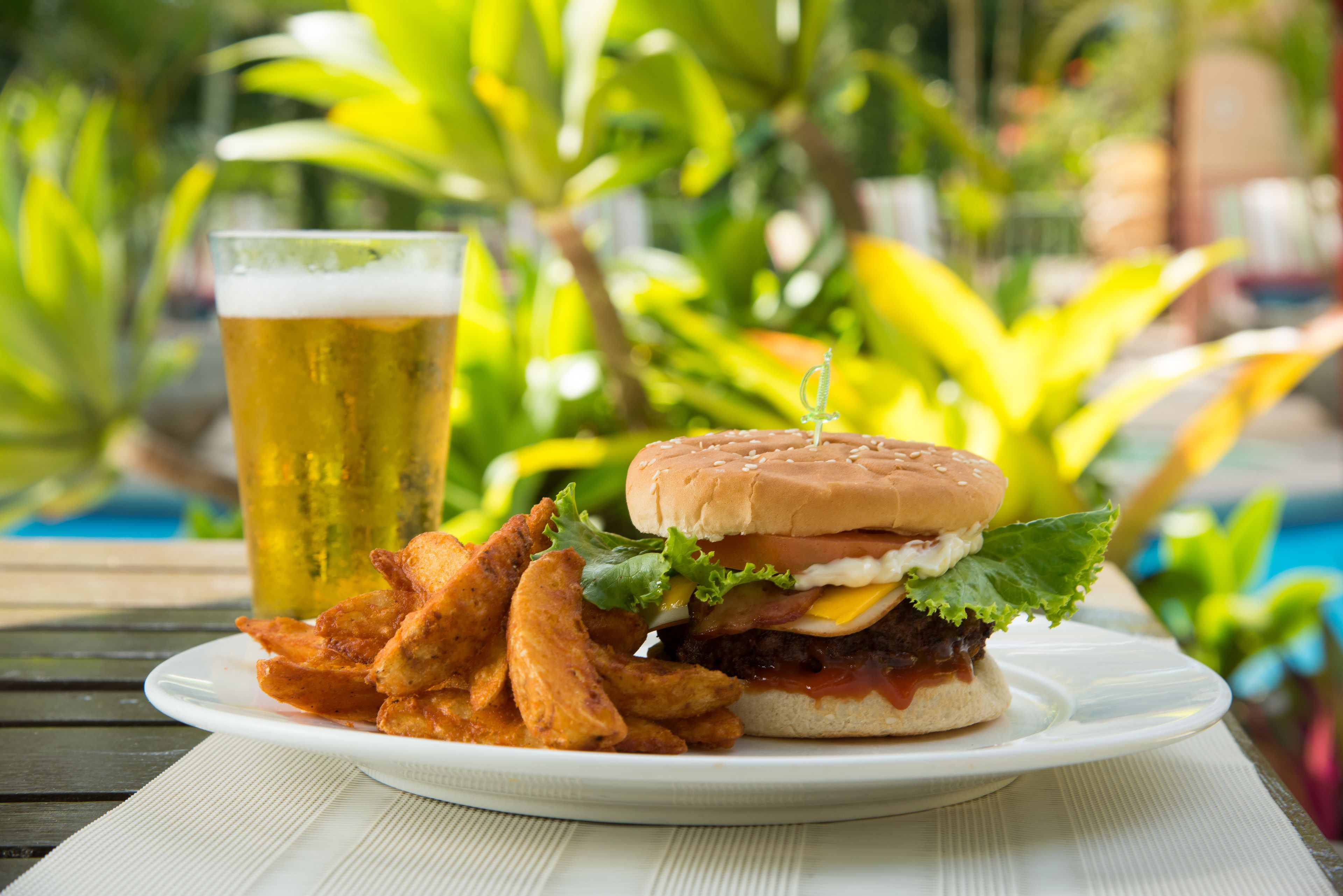 Ein Teller mit einem Burger und Pommes, dazu ein Glas Bier auf einem Tisch. Im Hintergrund ist ein Pool zu sehen.