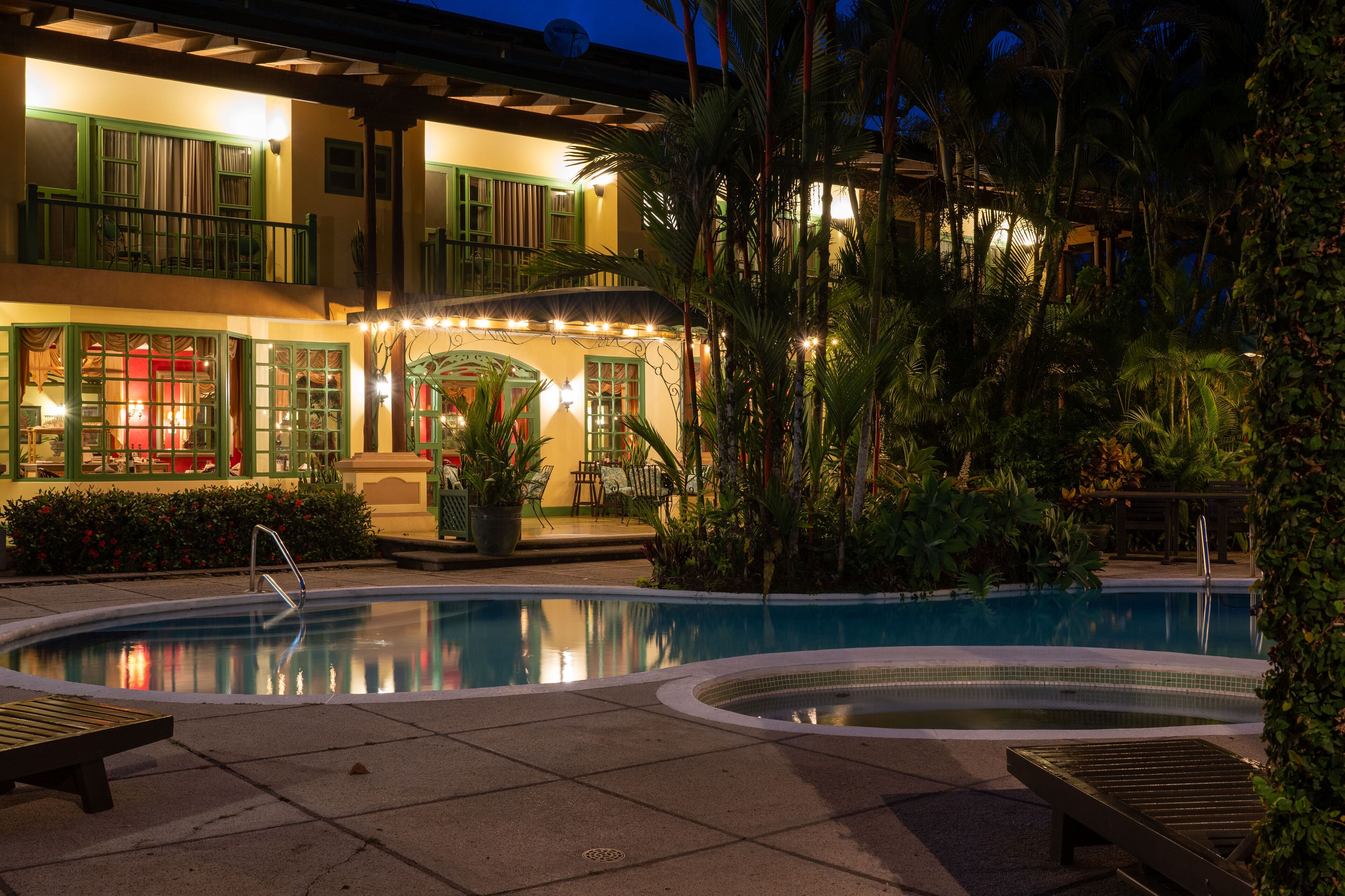 A pool area with lounge chairs, a hot tub, and a building with green shutters and outdoor seating.
