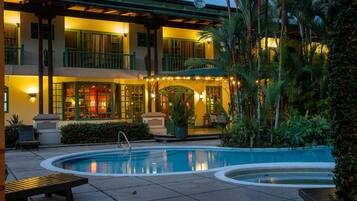 Zona de piscina con una silla de descanso, una piscina pequeña y un edificio con múltiples balcones e iluminación exterior.