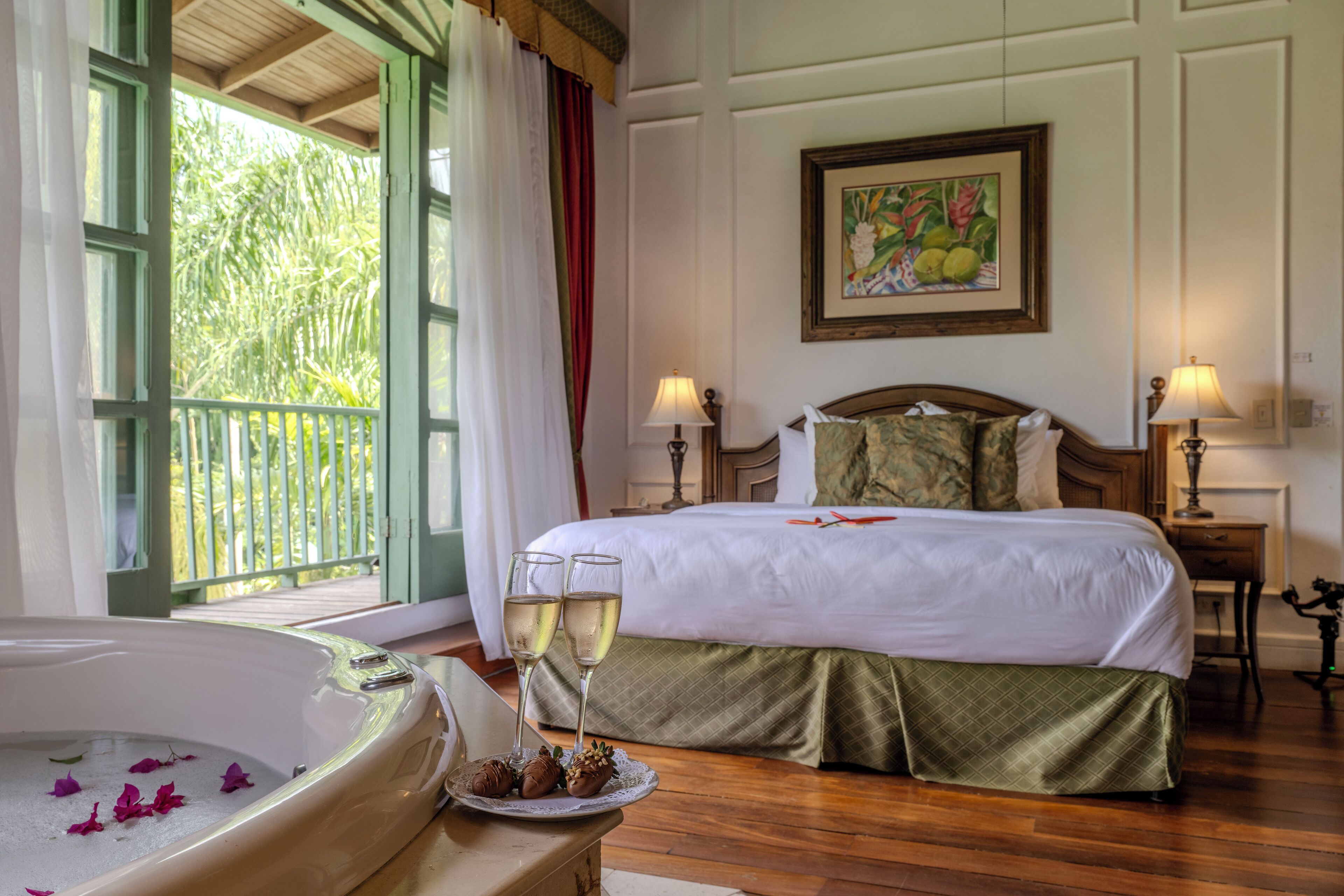 A hotel room with a bed, two bedside lamps, a painting on the wall, a window with curtains, and a bathtub with rose petals and a plate of food.