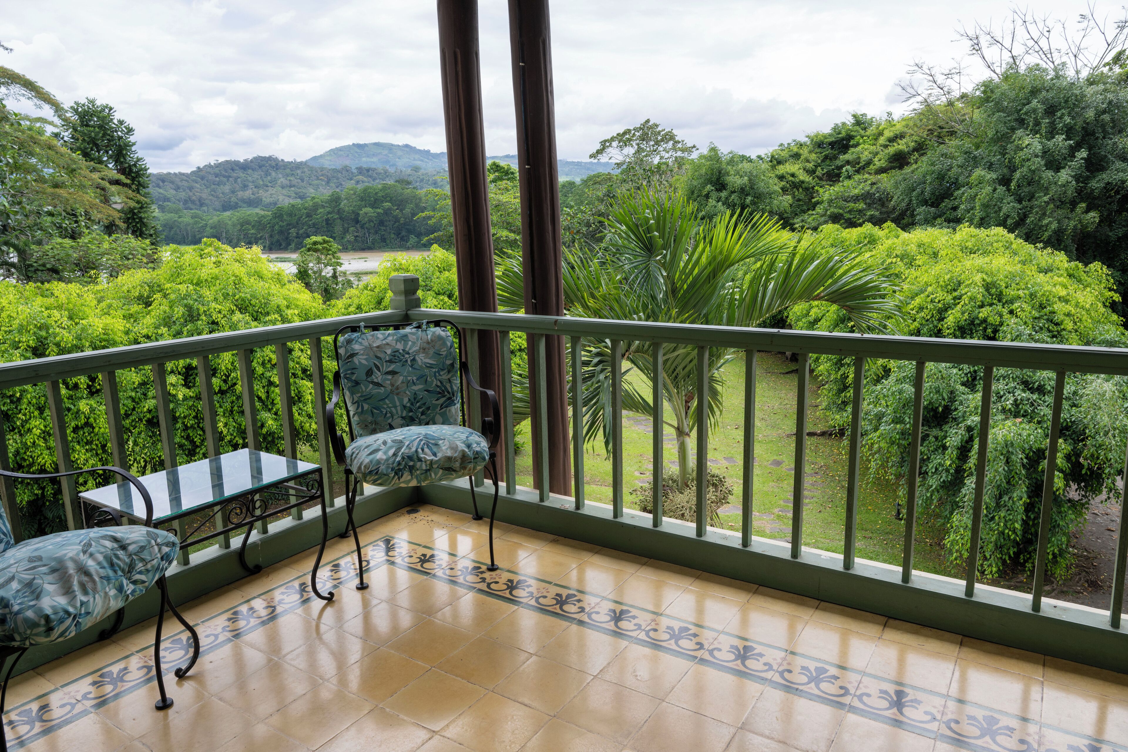 A balcony with a glass table and two chairs, overlooking a lush garden and distant hills.