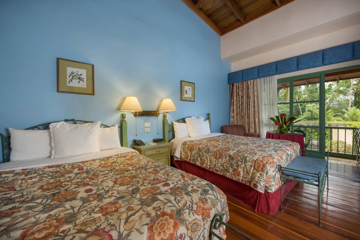 A room with two beds, a nightstand, a window with curtains, and a view of greenery outside.