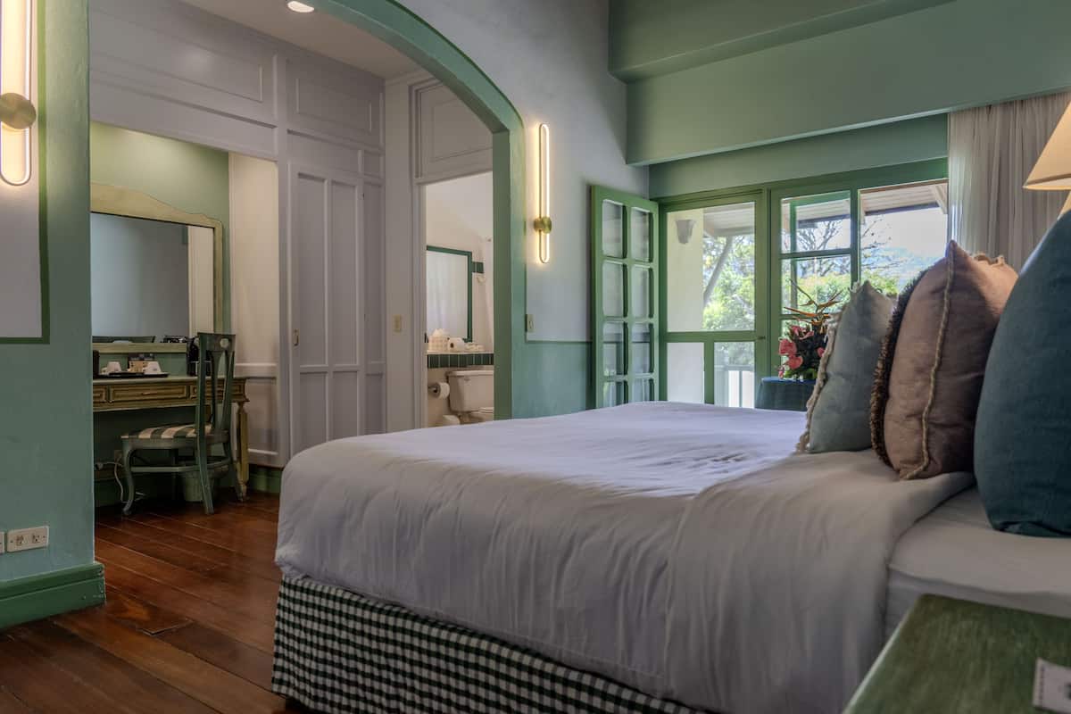 A bedroom with a large bed, a dresser, and a vanity with a mirror. There is a window with curtains and a view of greenery outside.