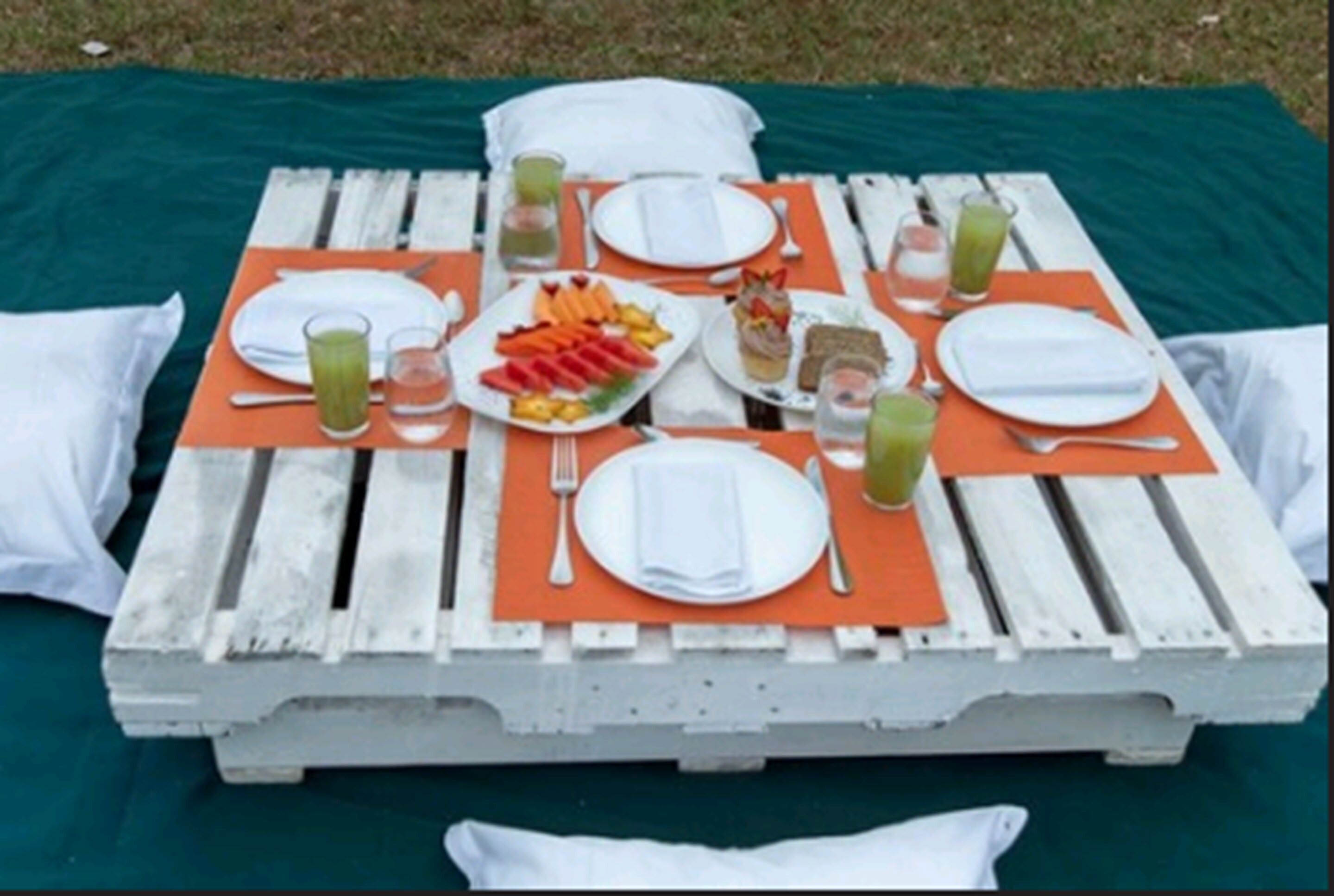 Ein Picknick auf einem Holztisch, mit orangefarbenen Tischsets, weißen Tellern und verschiedenen Speisen, darunter Obst und Getränke.