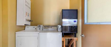 A laundry area with a washer and dryer, a shelf with a black appliance, and a whiteboard with cleaning supplies.