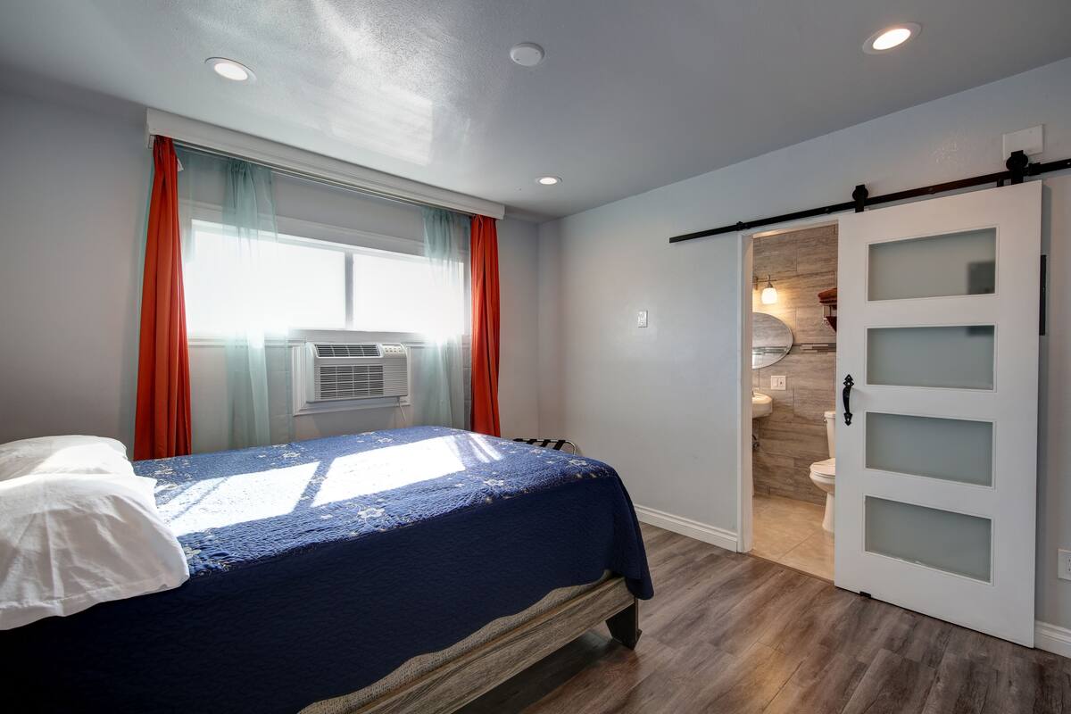 Un dormitorio con cama, una puerta corredera al baño y una ventana con cortinas.
