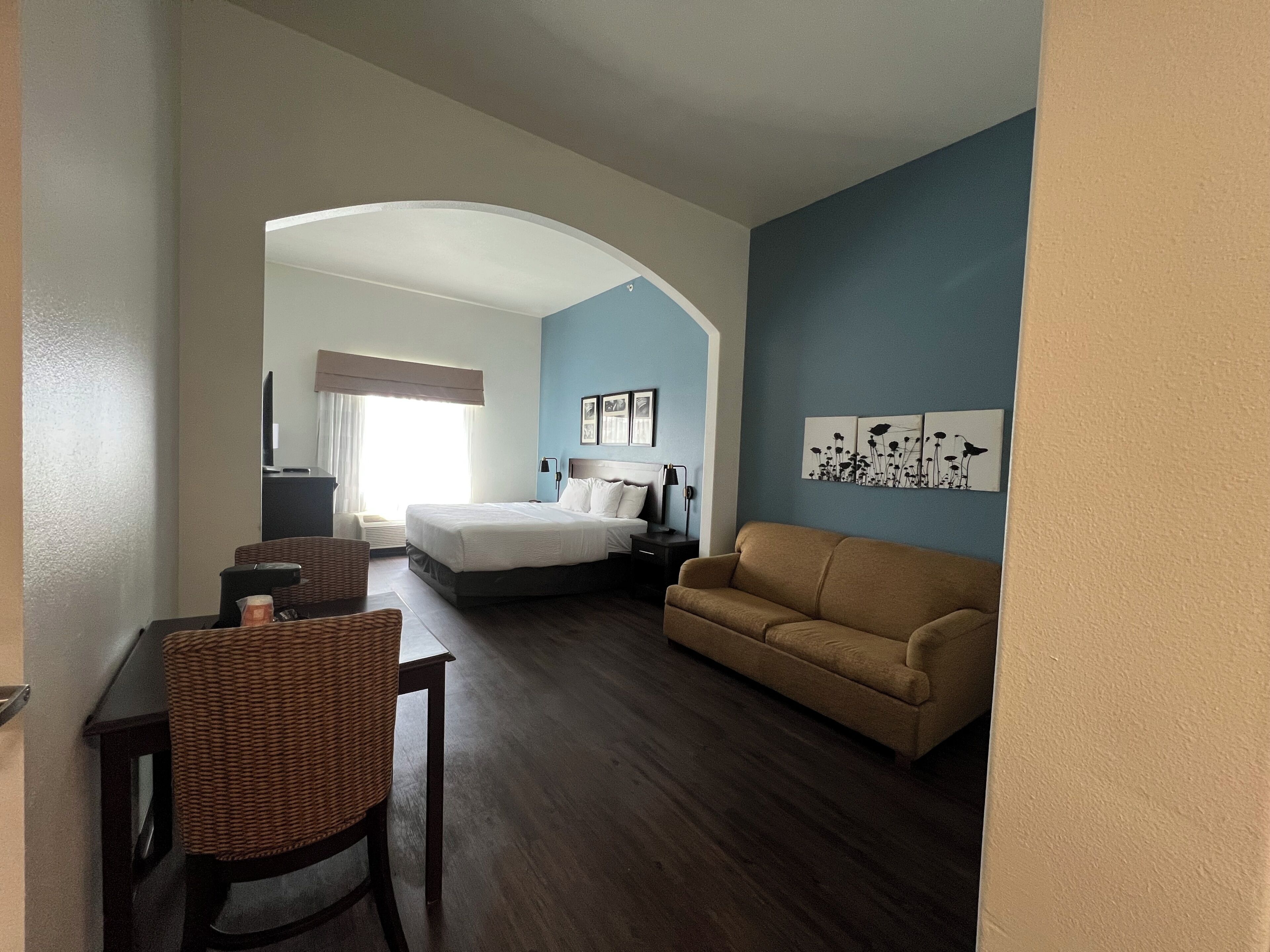 A hotel room with a bed, a sofa, a desk with a chair, and artwork on the wall.