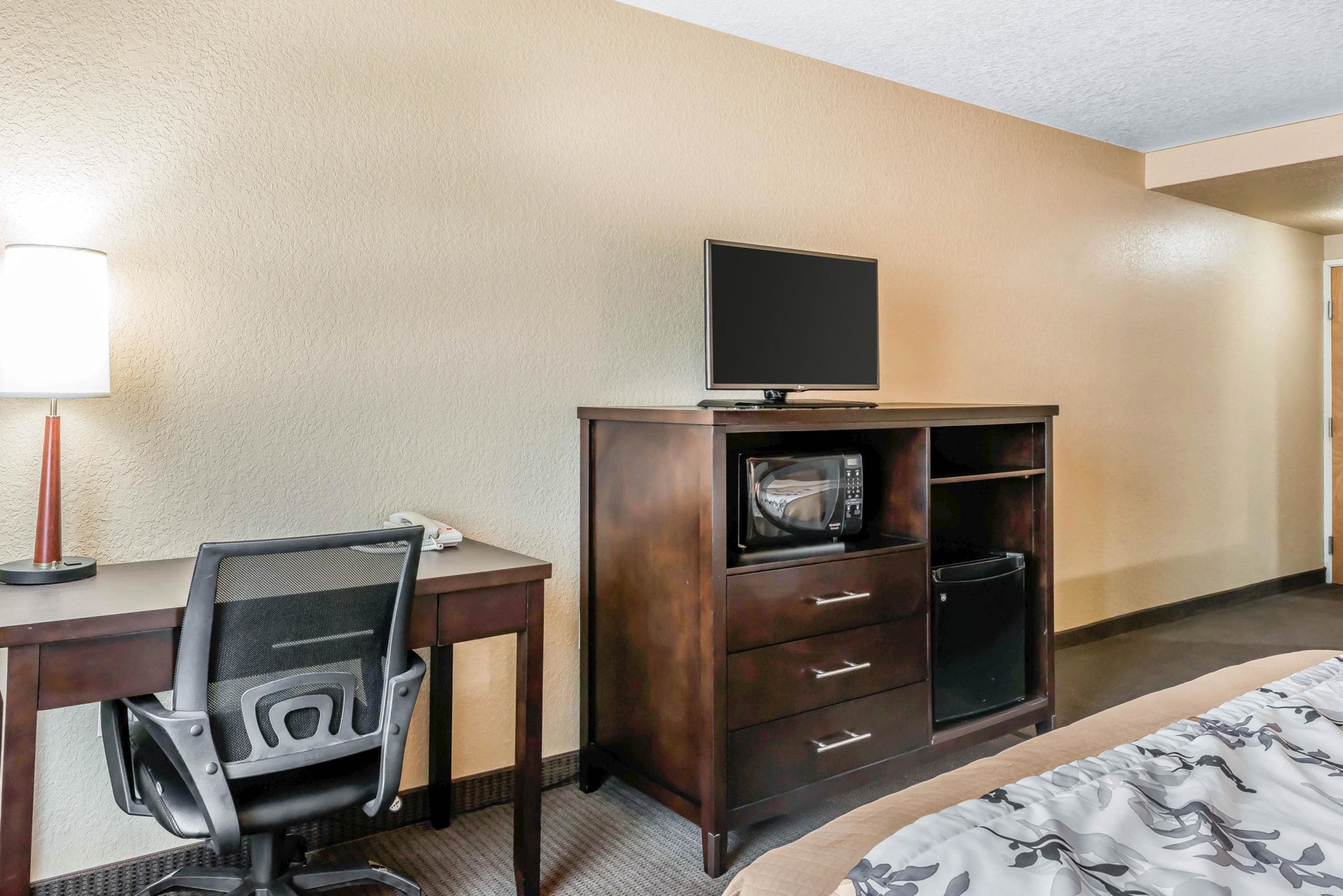 A hotel room with a desk, a chair, a television on a cabinet, a microwave, and a bed.