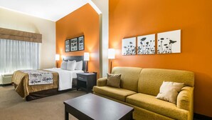 A hotel room with a bed, a sofa, a coffee table, and wall art. - Sleep Inn and Suites - Ocala / Belleview (Ocala)