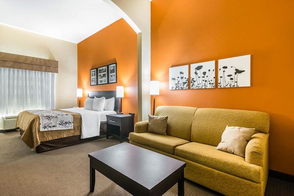 A hotel room with a bed, a sofa, a coffee table, and wall art. - Sleep Inn and Suites - Ocala / Belleview (Ocala)