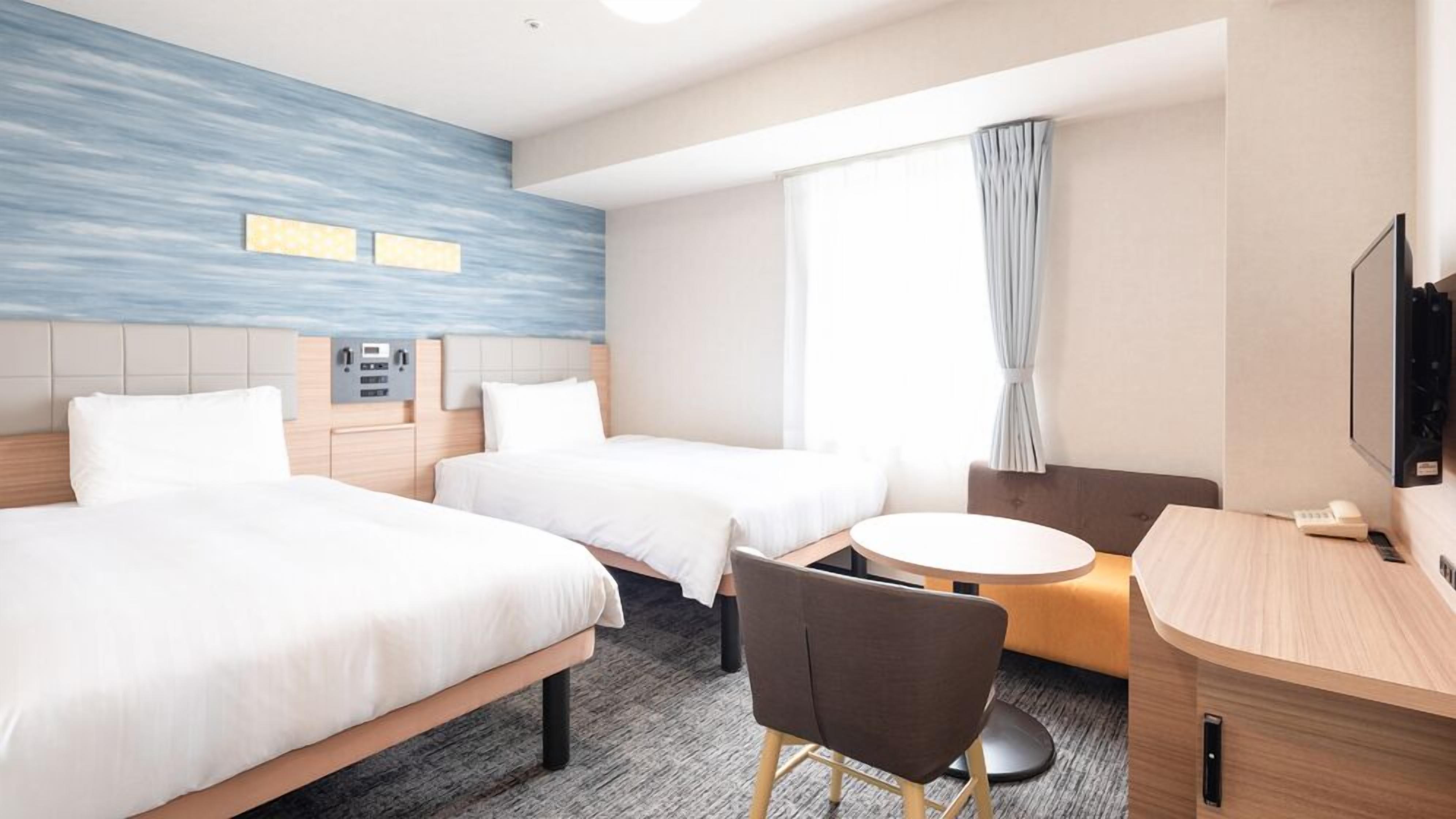A hotel room with two beds, a desk, a chair, a small round table, a TV, and a window with curtains.
