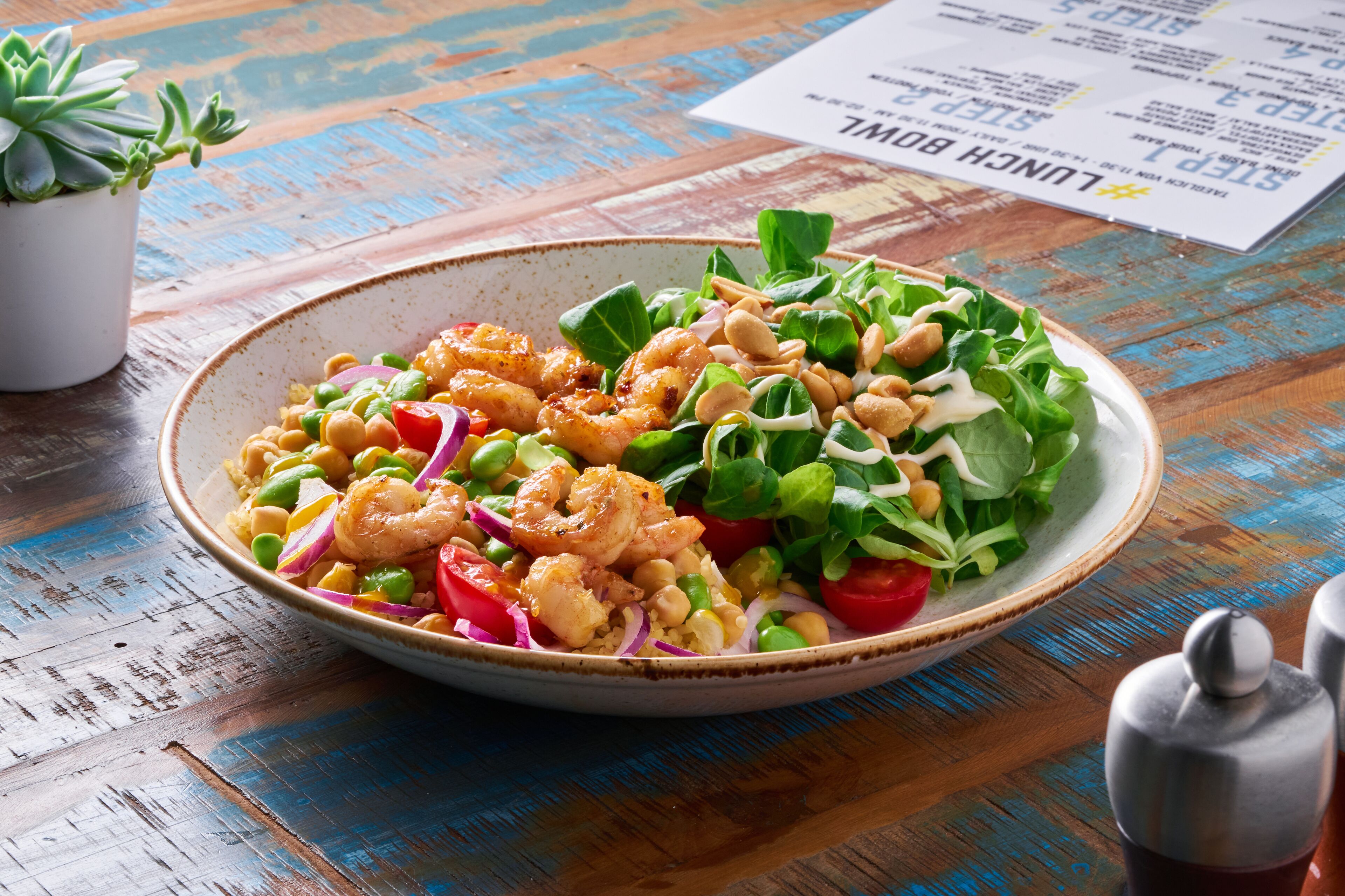 Uma tigela de camarão e salada sobre uma mesa de madeira, com um menu ao fundo.