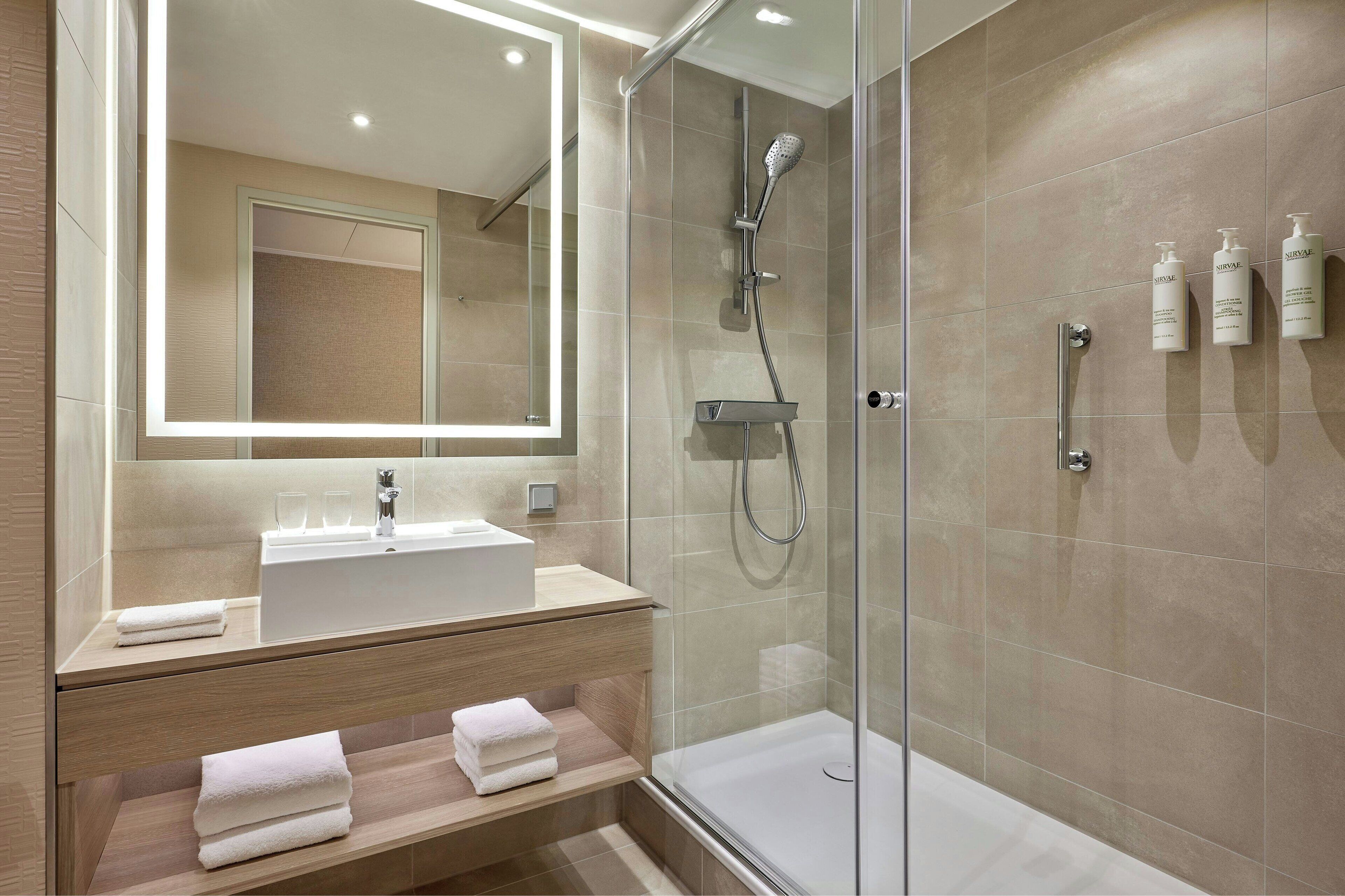 Un baño moderno con ducha envuelta en vidrio, un espejo grande y un tocador con lavamanos y toallero.