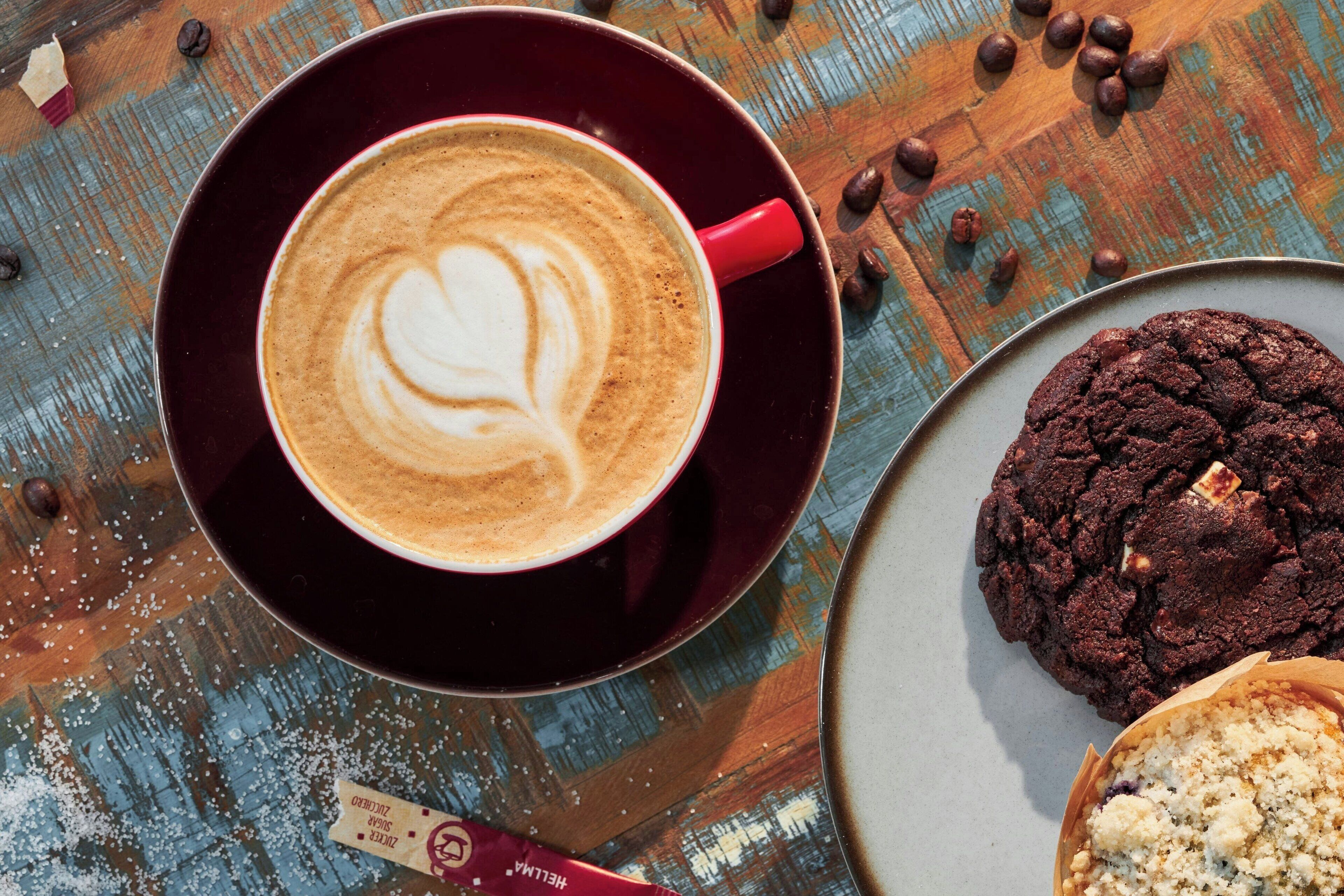Una taza de café con arte de latte sobre un plato, una galleta de chocolate y un bizcocho en un plato.