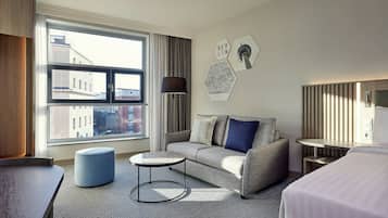 A modern hotel room with a sofa, a round coffee table, a bed, and a view of buildings through large windows.