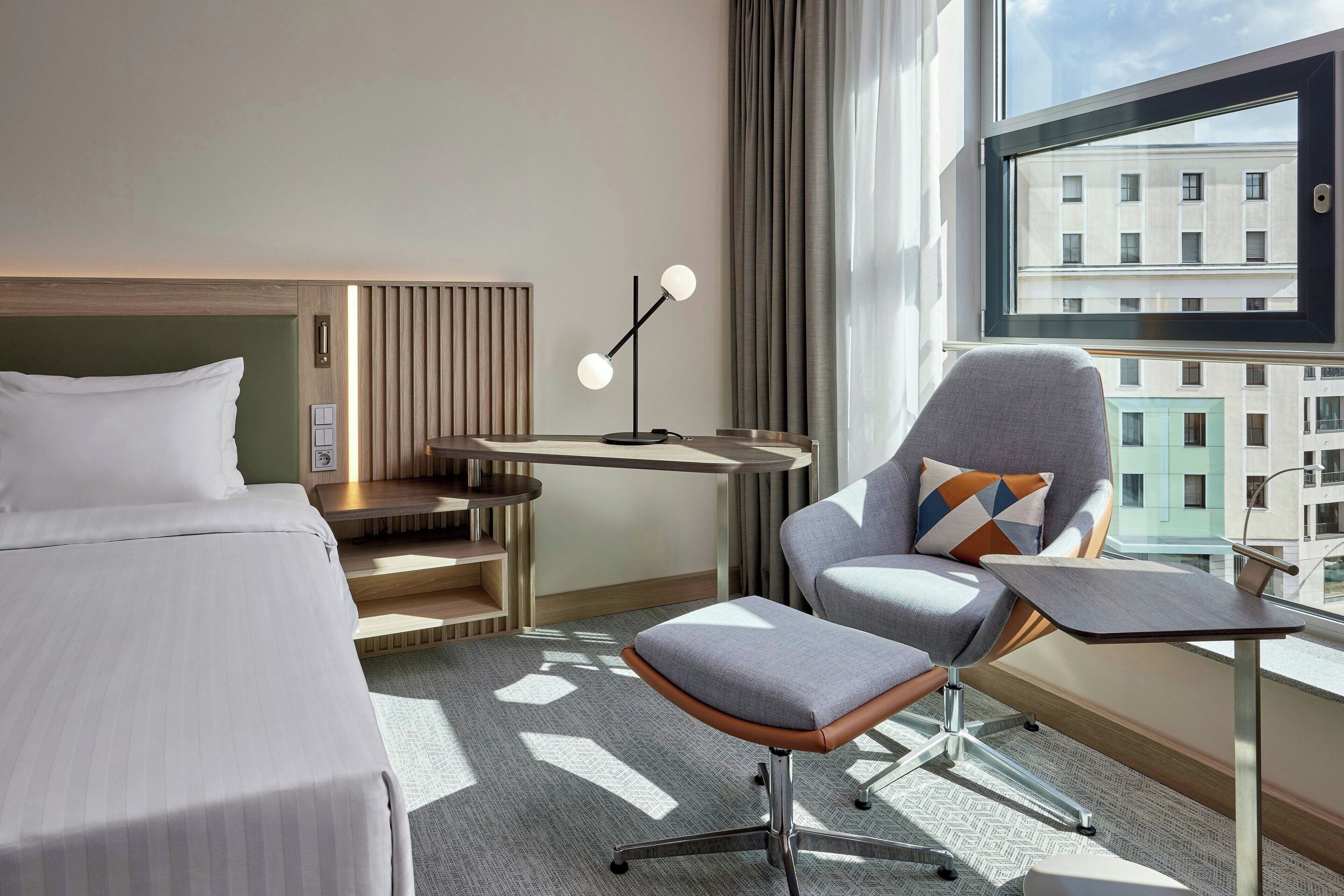 A hotel room with a bed, a desk, a chair, and a view of buildings through a window.