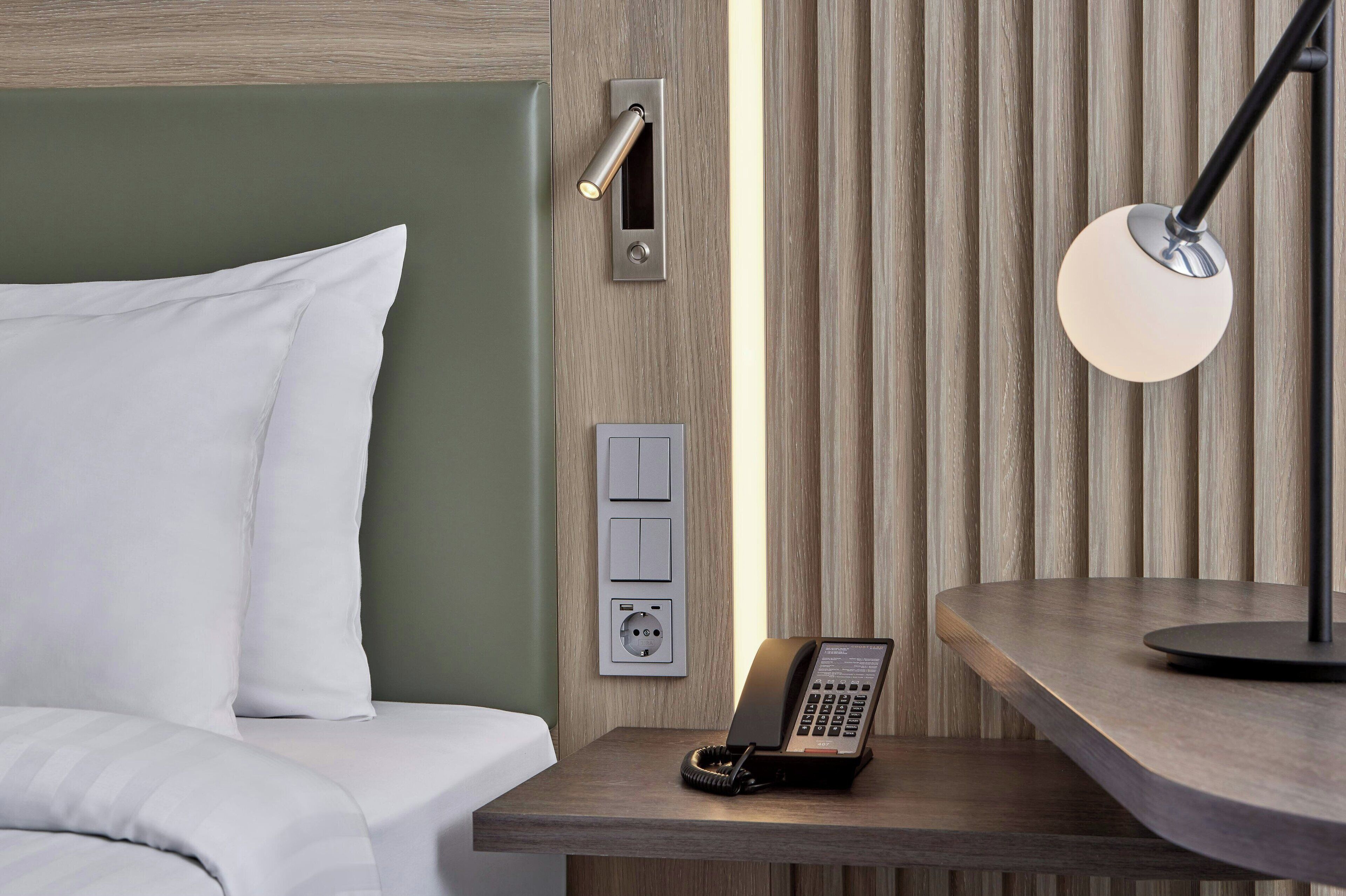 A hotel room with a bed, a bedside telephone, a wall socket, and a bedside lamp.