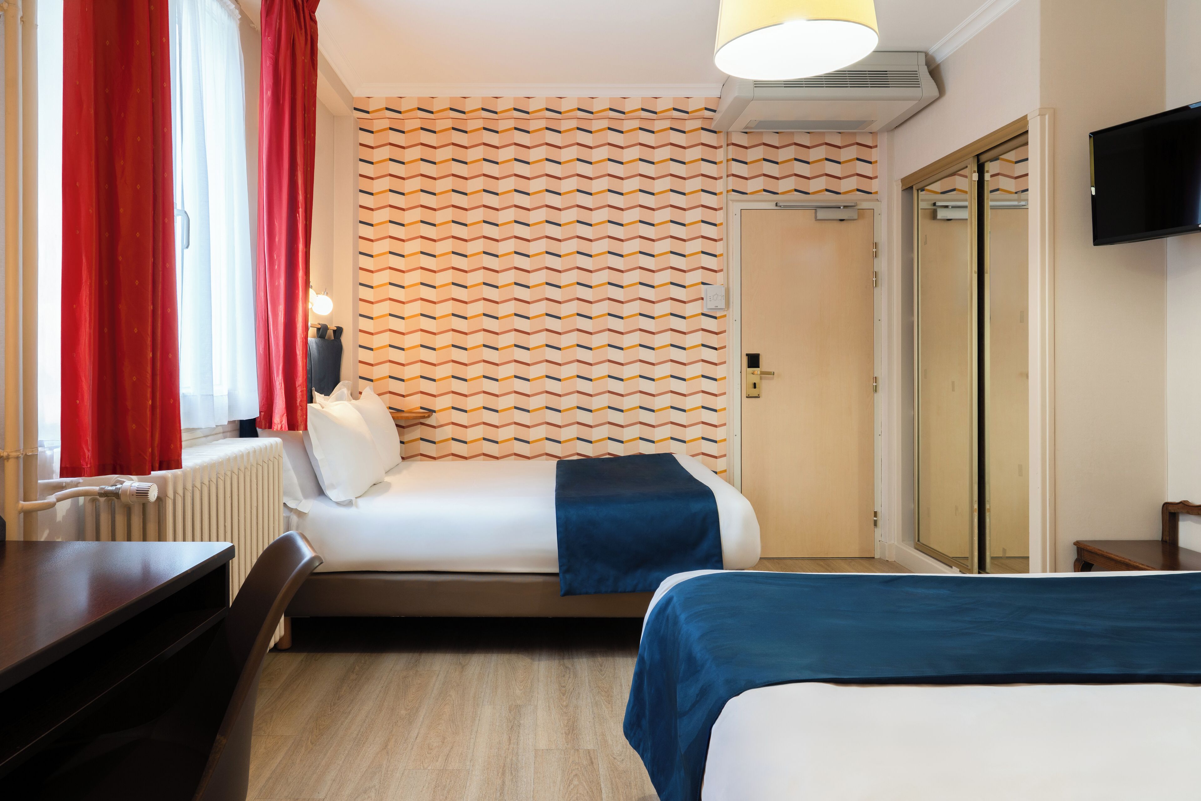A hotel room with a bed, a desk, a chair, a radiator, a television, and a door with a mirror.